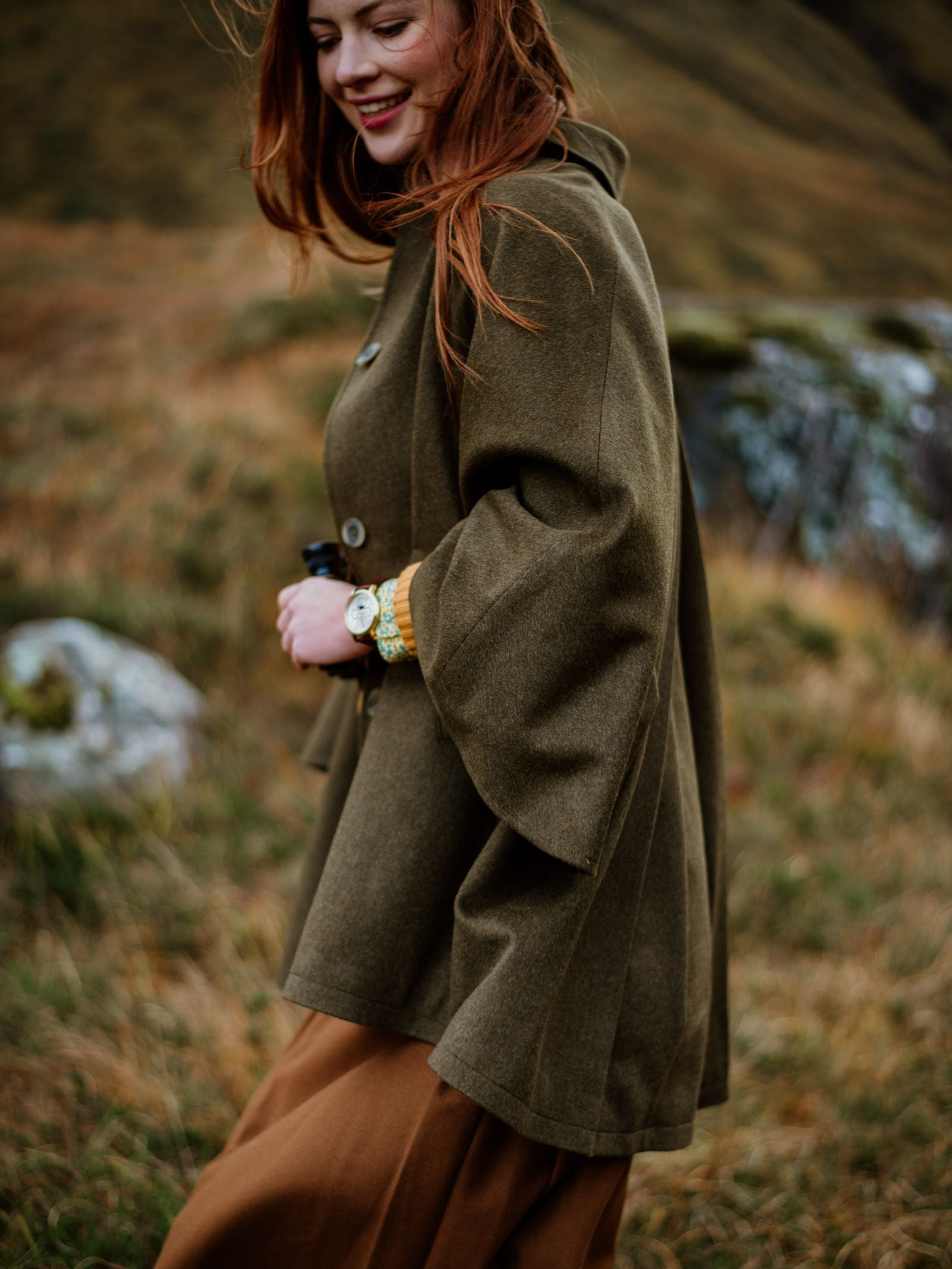 A woman with long auburn hair smiles outdoors in a grassy, hilly landscape, wearing the Campbell's of Beauly Loden Green Cape and a brown skirt. She holds something in her hand and wears a wristwatch.