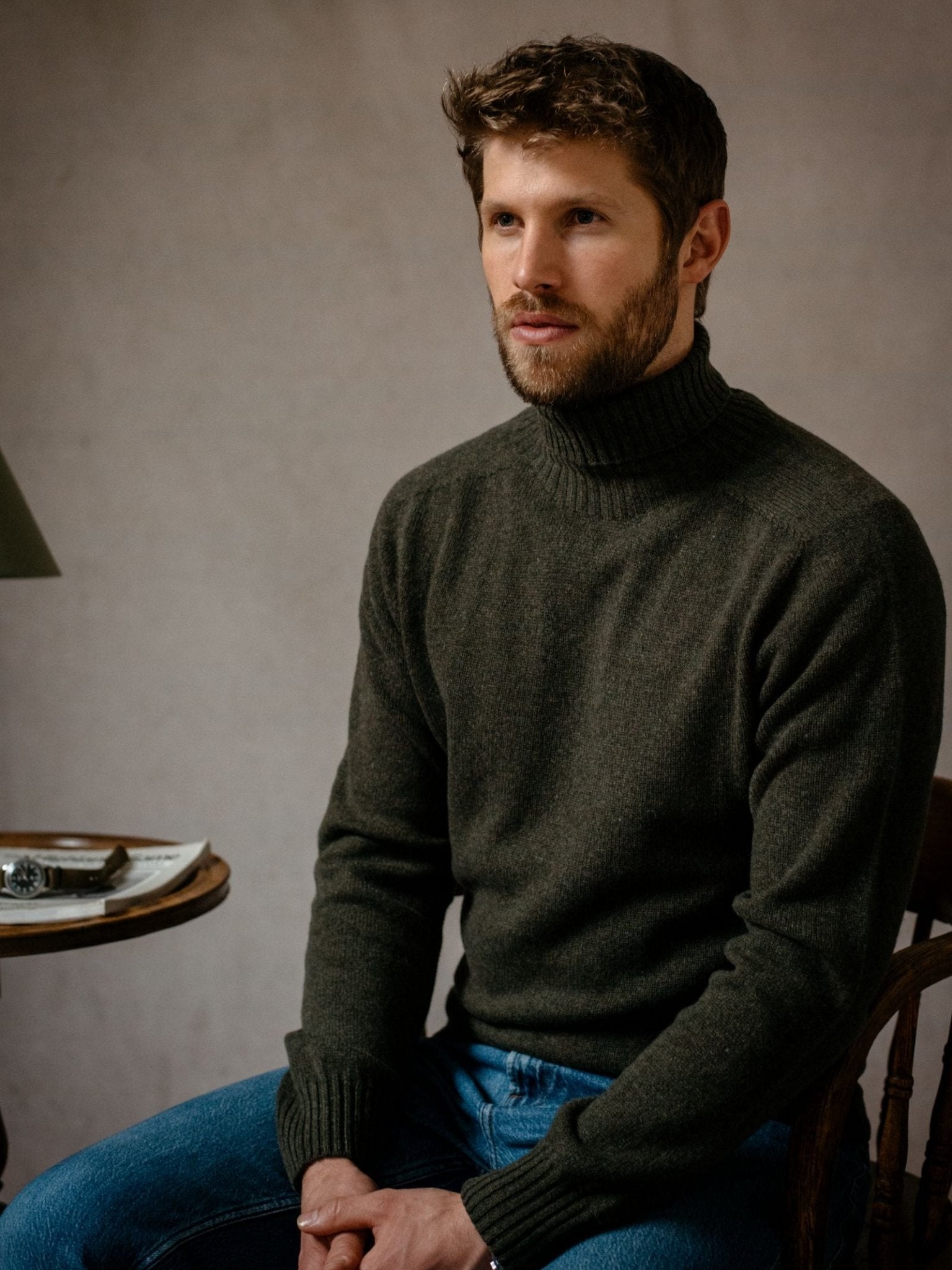 A man with short brown hair and a beard sits on a wooden chair, wearing the Campbell's of Beauly Lambswool Roll Neck in dark green with blue jeans. He looks slightly to the side beside a small table holding a lamp and book.