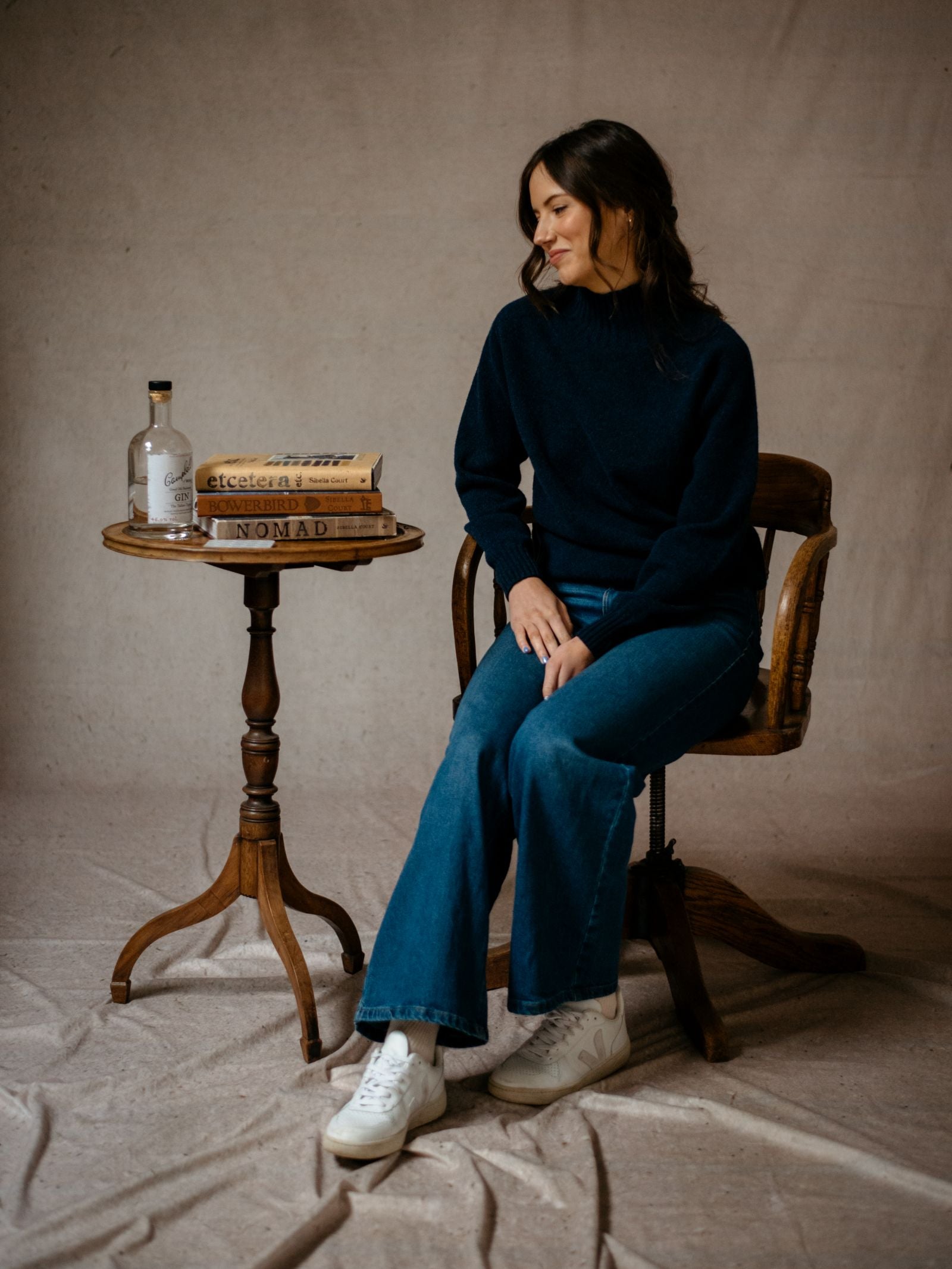 A woman in a navy Campbells of Beauly Geelong Lambswool Funnel Neck sweater and jeans sits on a wooden chair, smiling to the side beside a small round table with books and a clear bottle, set against a neutral backdrop.