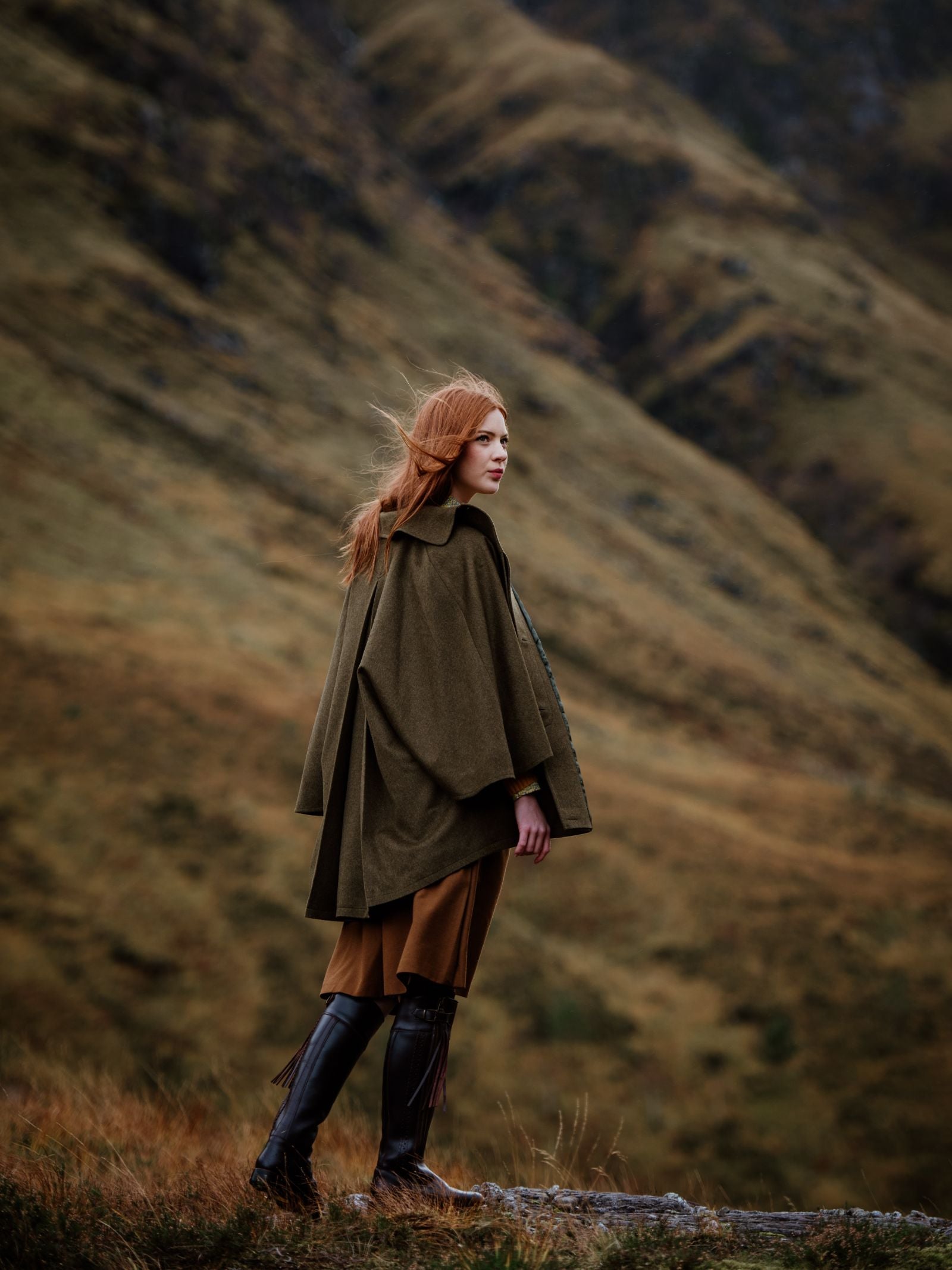 A woman with long red hair in an olive-green cape, brown pants, and Campbells of Beauly Leather Spanish Boots with a Vibram outsole stands on grassy terrain before windswept hills and mountains, evoking a rugged, natural atmosphere.
