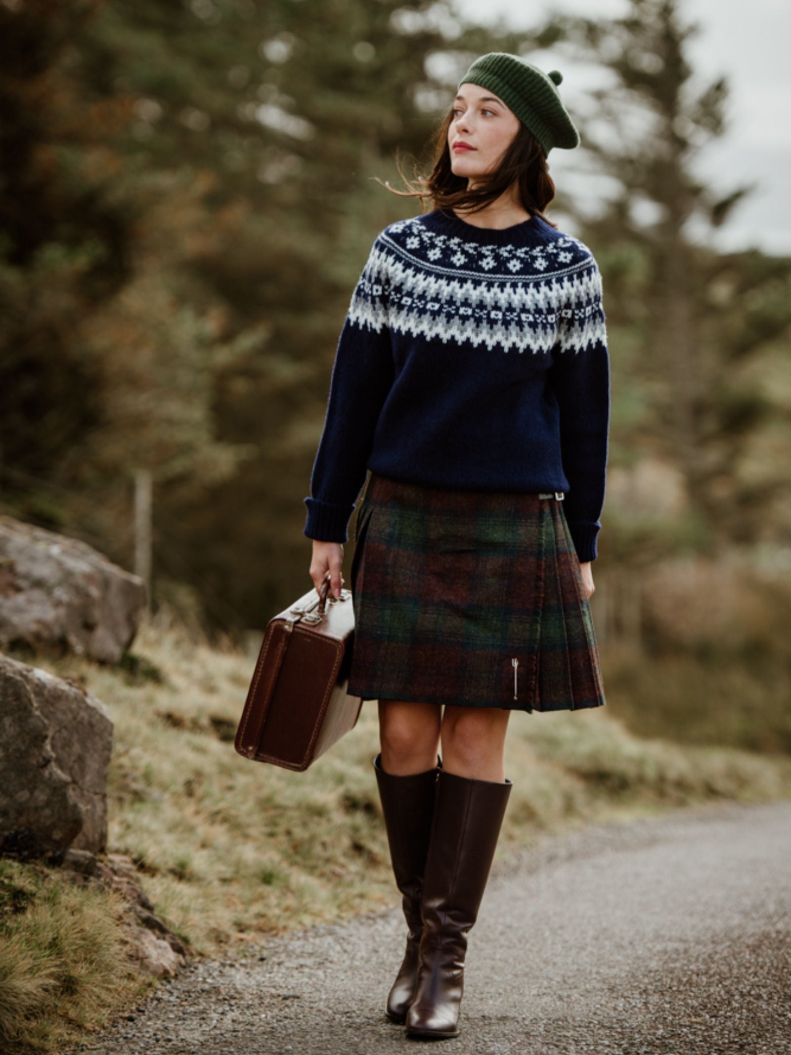 A woman in Campbell's of Beauly Rib Beret, navy patterned sweater, plaid skirt, and brown boots walks outdoors with a brown suitcase—an effortlessly stylish winter look among trees and grass.