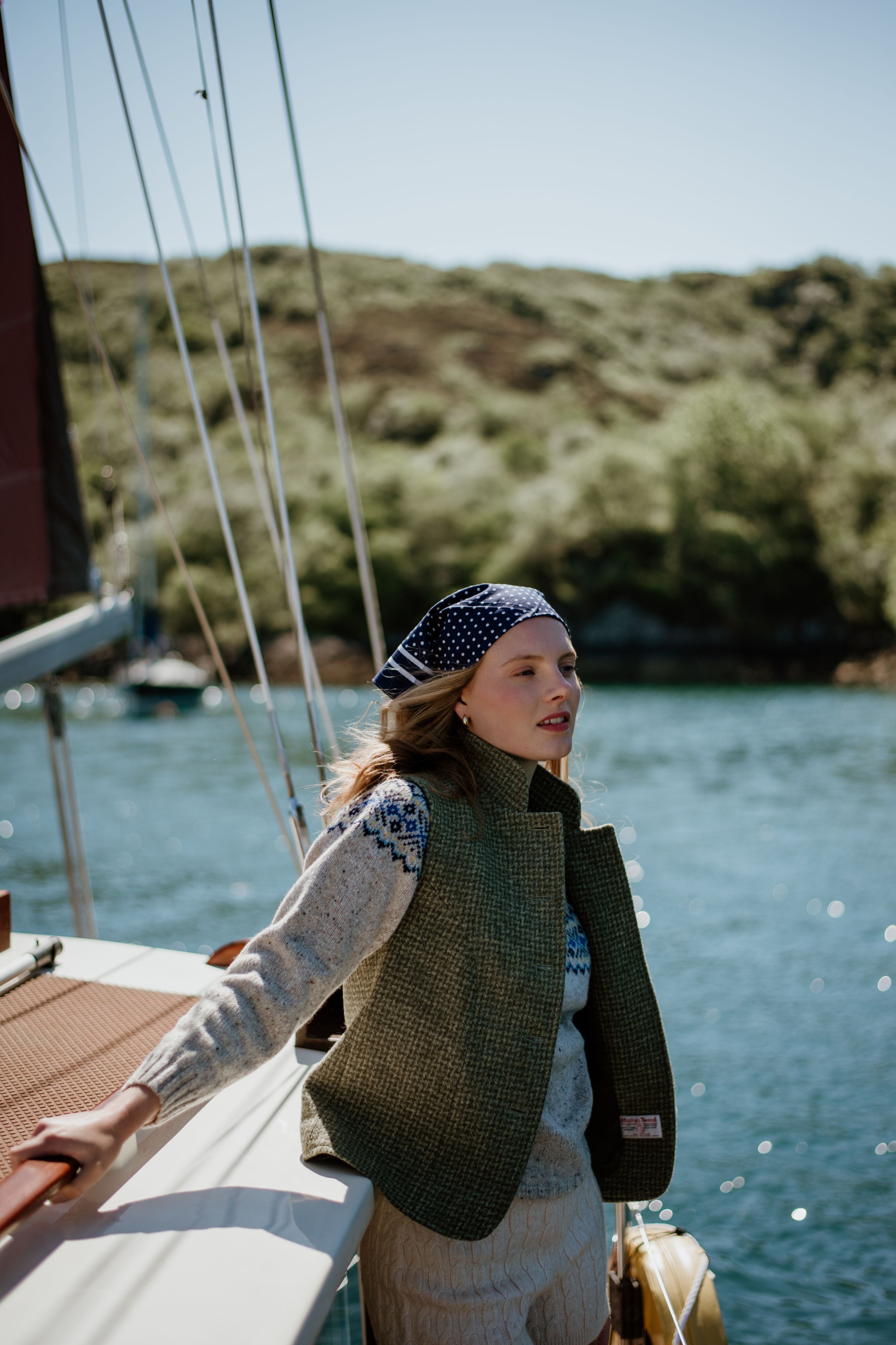A woman wearing a patterned headscarf, knitted sweater, and vest stands on a sailboat, holding onto the rail, with water and a green, hilly shoreline in the background under a clear sky.