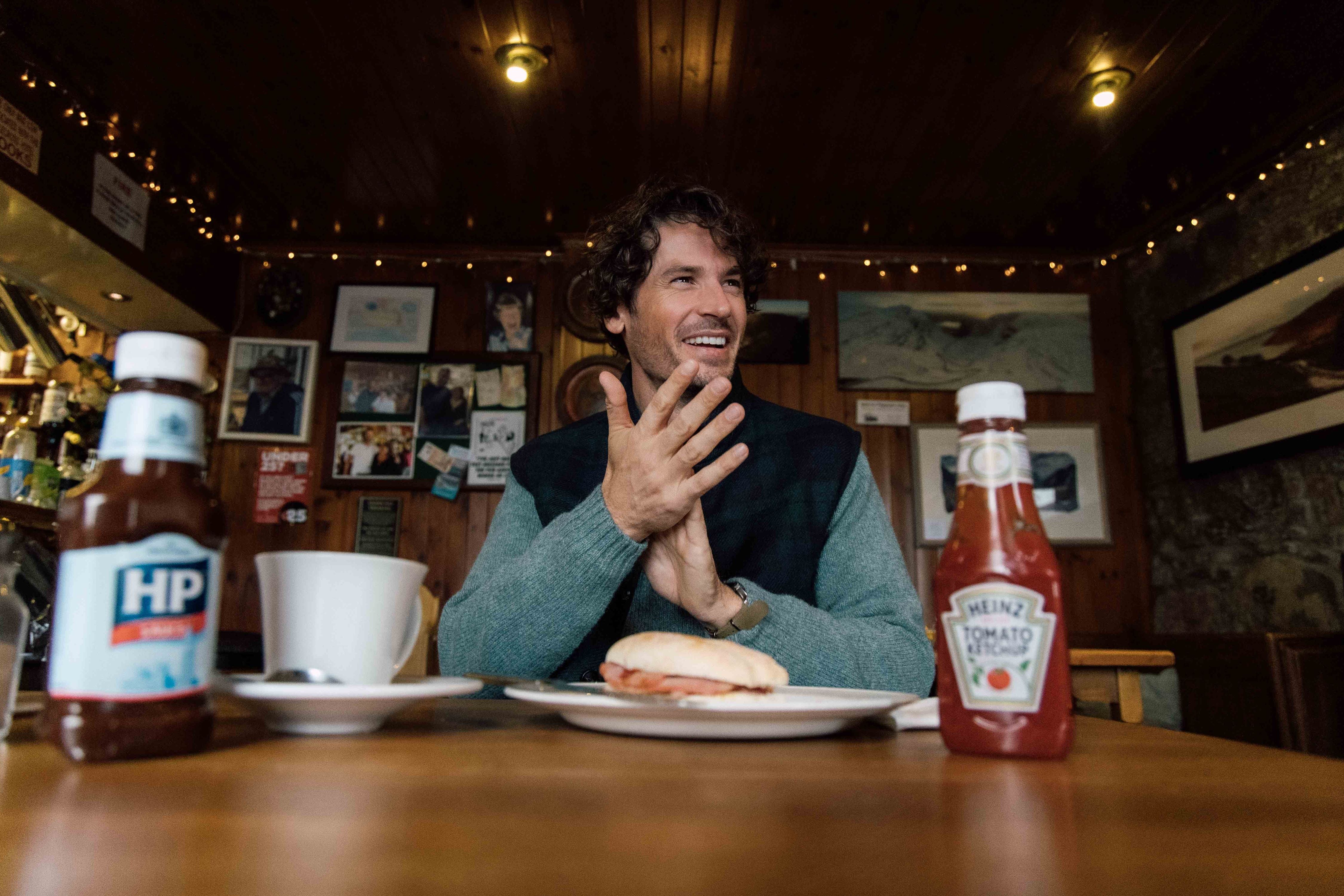 A man sits at a wooden table in a cozy cafe, smiling with his hands together. In front of him are a plate with a sandwich, a white mug, and bottles of HP Sauce and Heinz Ketchup. Warm lights and pictures decorate the background.