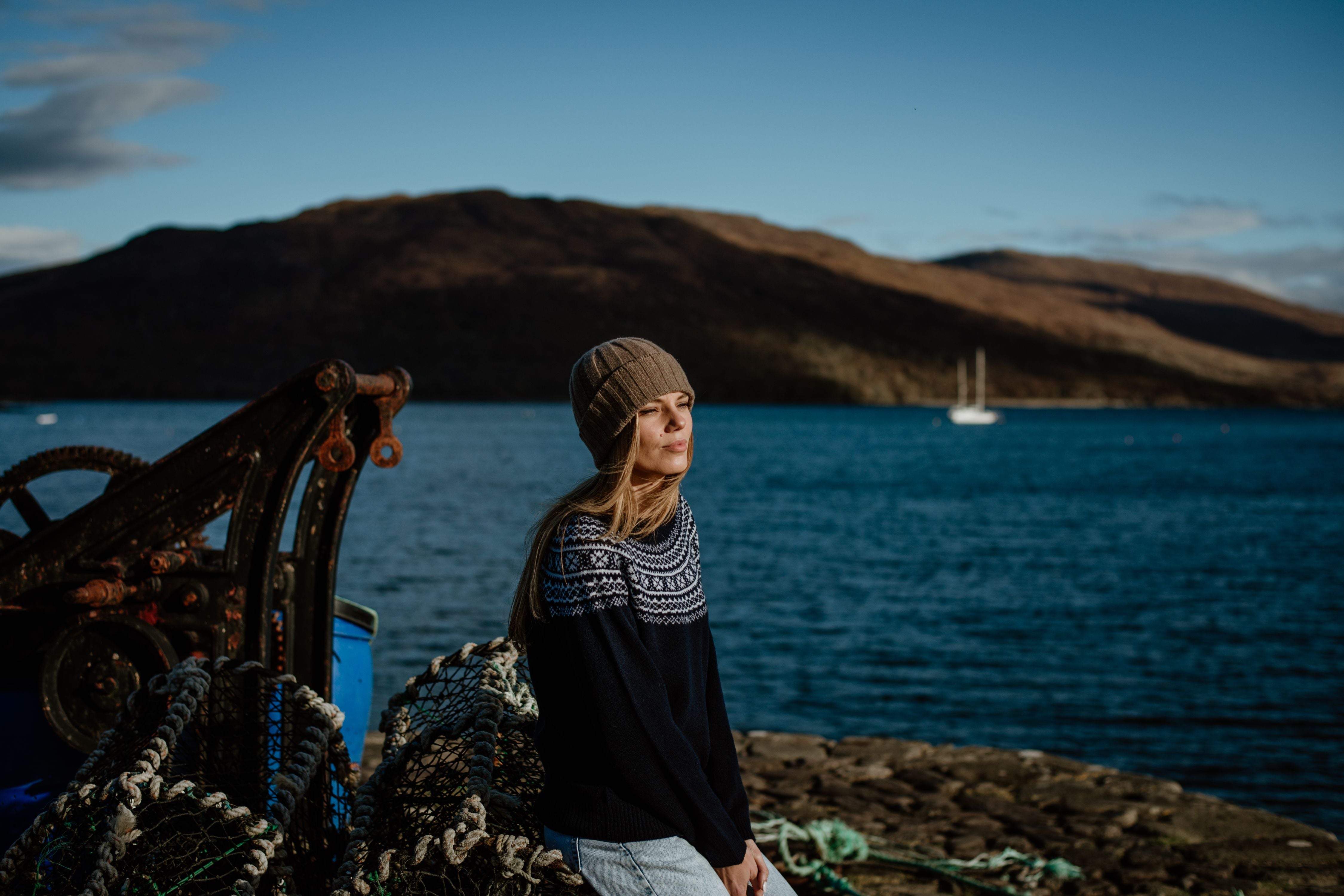 A woman in a knit hat and sweater sits by fishing nets near the edge of a body of water, with hills and a sailboat in the background under a clear blue sky.
