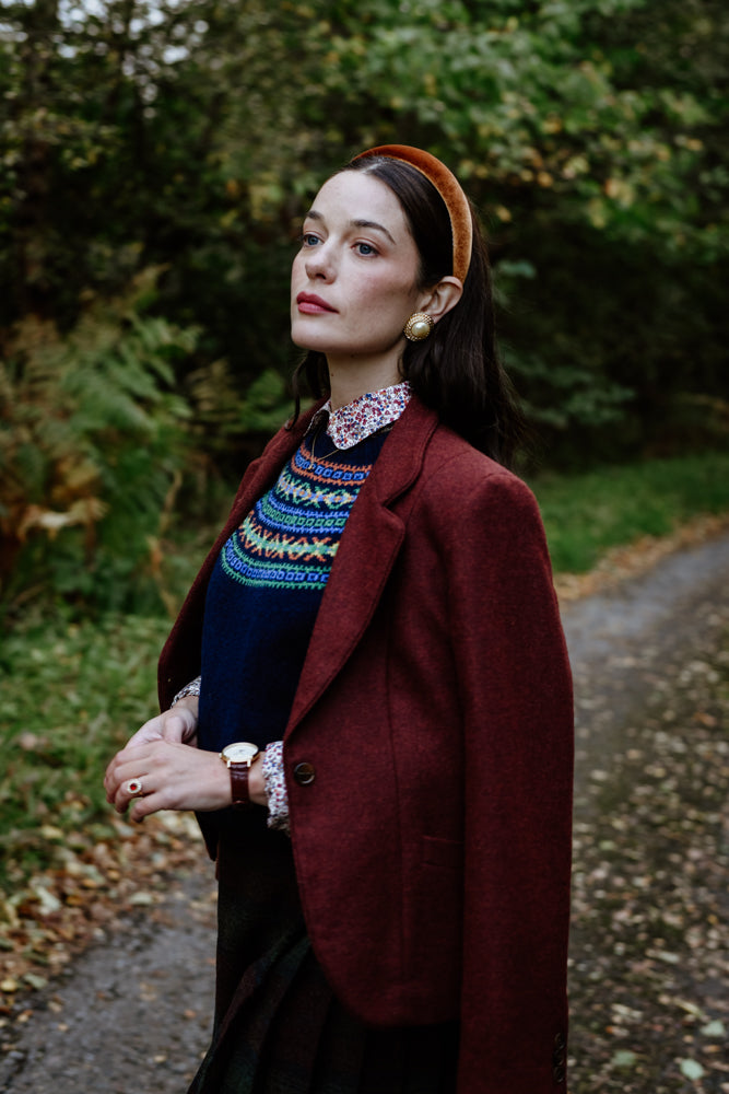 A woman wearing a burgundy blazer, patterned sweater, floral shirt, and headband stands on a leaf-covered path in a green, wooded outdoor setting, looking thoughtful.