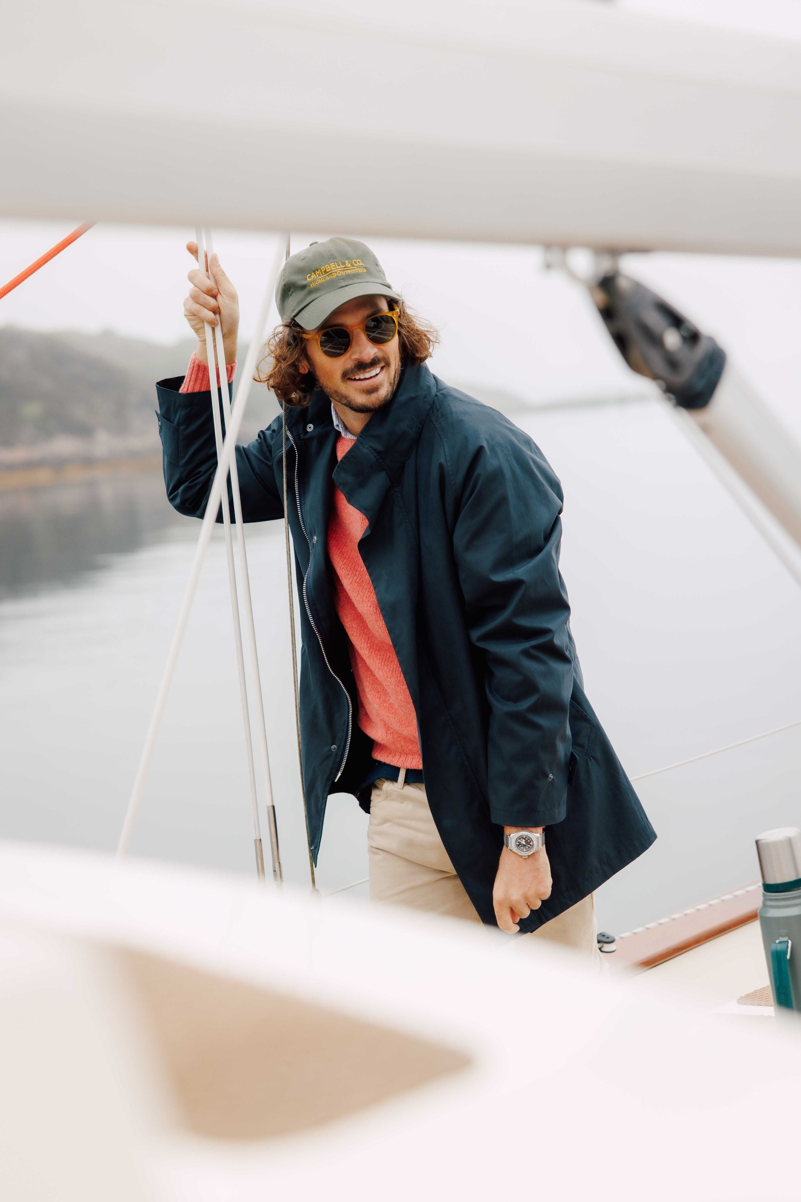 A man wearing a green cap, sunglasses, a navy jacket, red sweater, and beige pants stands on a sailboat, holding a rope and smiling. The background shows calm water and a blurred shoreline.