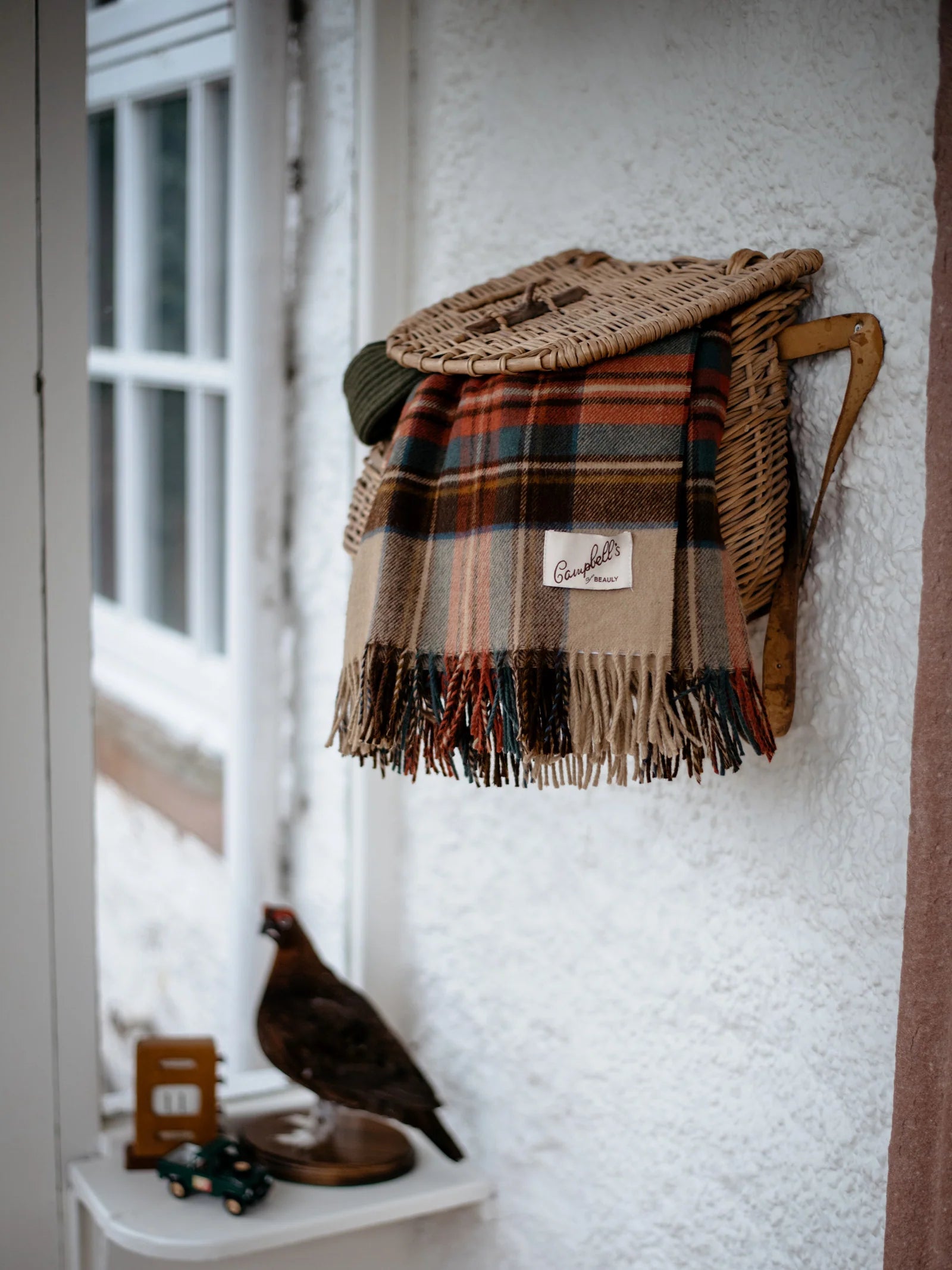 A Campbell's of Beauly Lambswool Tartan Blanket with fringe hangs on a wicker rack against white stucco. Below, a shelf holds a pheasant figurine, miniature green truck, and a decorative calendar.