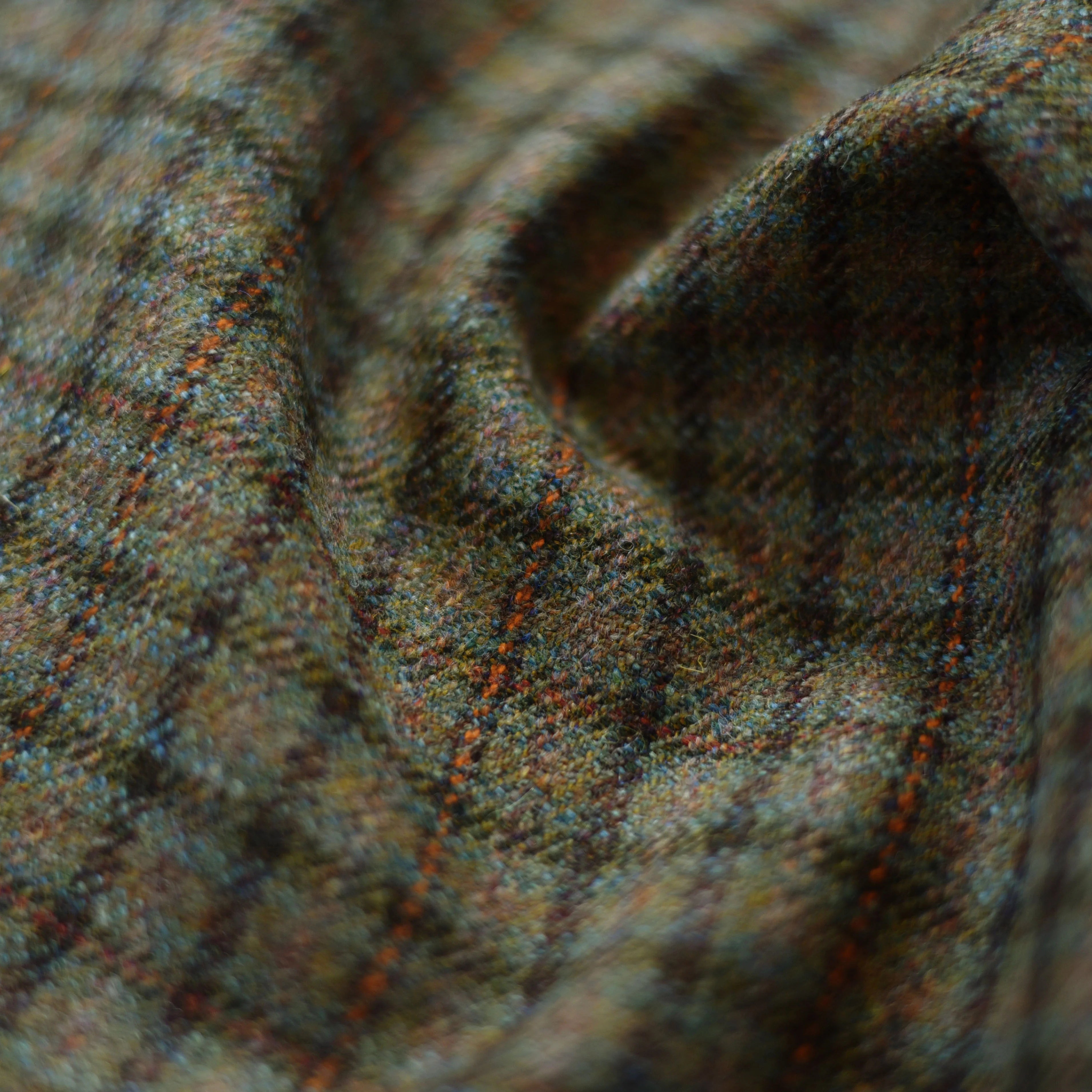 Close-up of House Tweed 71972 by Campbell's of Beauly, showcasing multicolored plaid in earthy tones with intersecting orange, green, and blue lines. The softly draped Scottish tweed forms gentle folds and shadows.