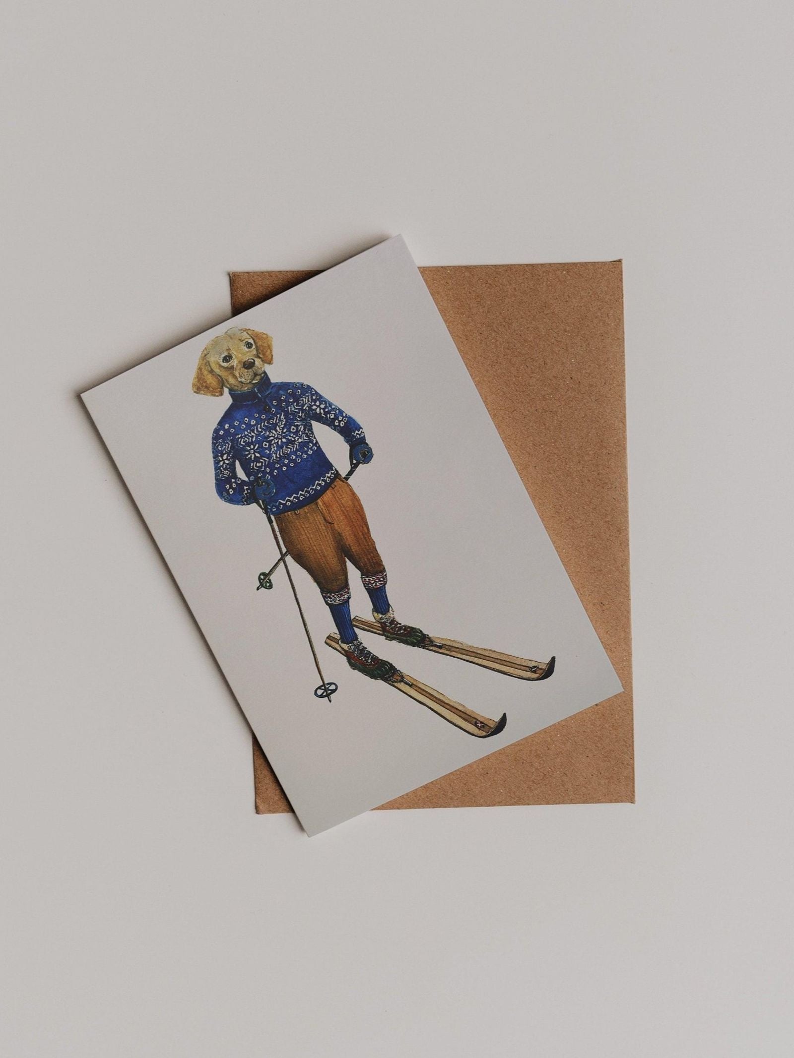 The Alpine Highlander Greeting Card by Campbell’s of Beauly features a charming illustration of a dog dressed in a blue sweater and brown pants, standing on skis with ski poles. Designed by Sophie Murray, the card is shown atop a brown envelope on a white surface.