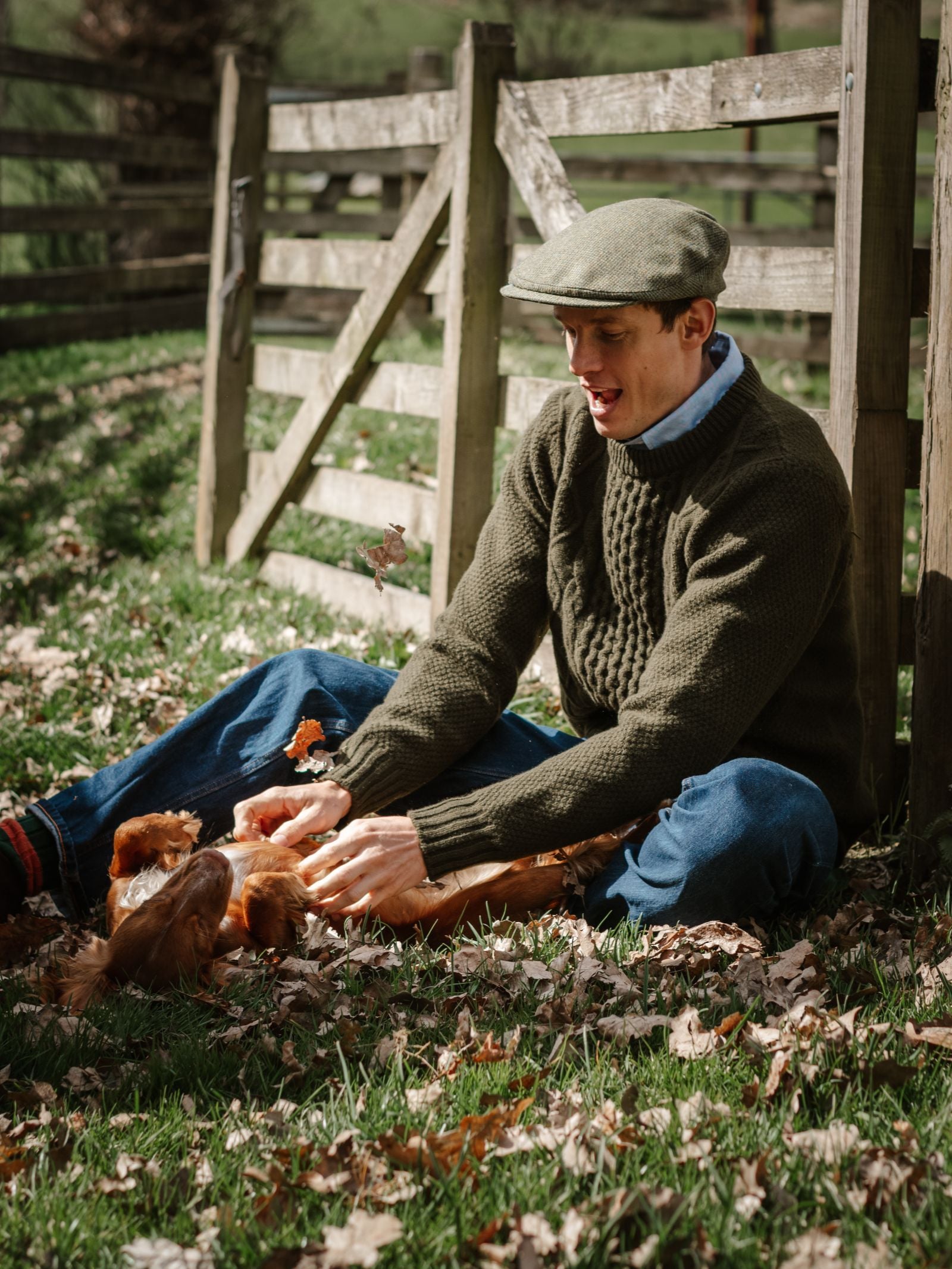 A man in a green sweater and the Campbell's of Beauly Unisex Beauly Cap in Green Melange sits on grass by a wooden fence, smiling and playing with brown chickens among scattered fallen leaves on a sunny day.