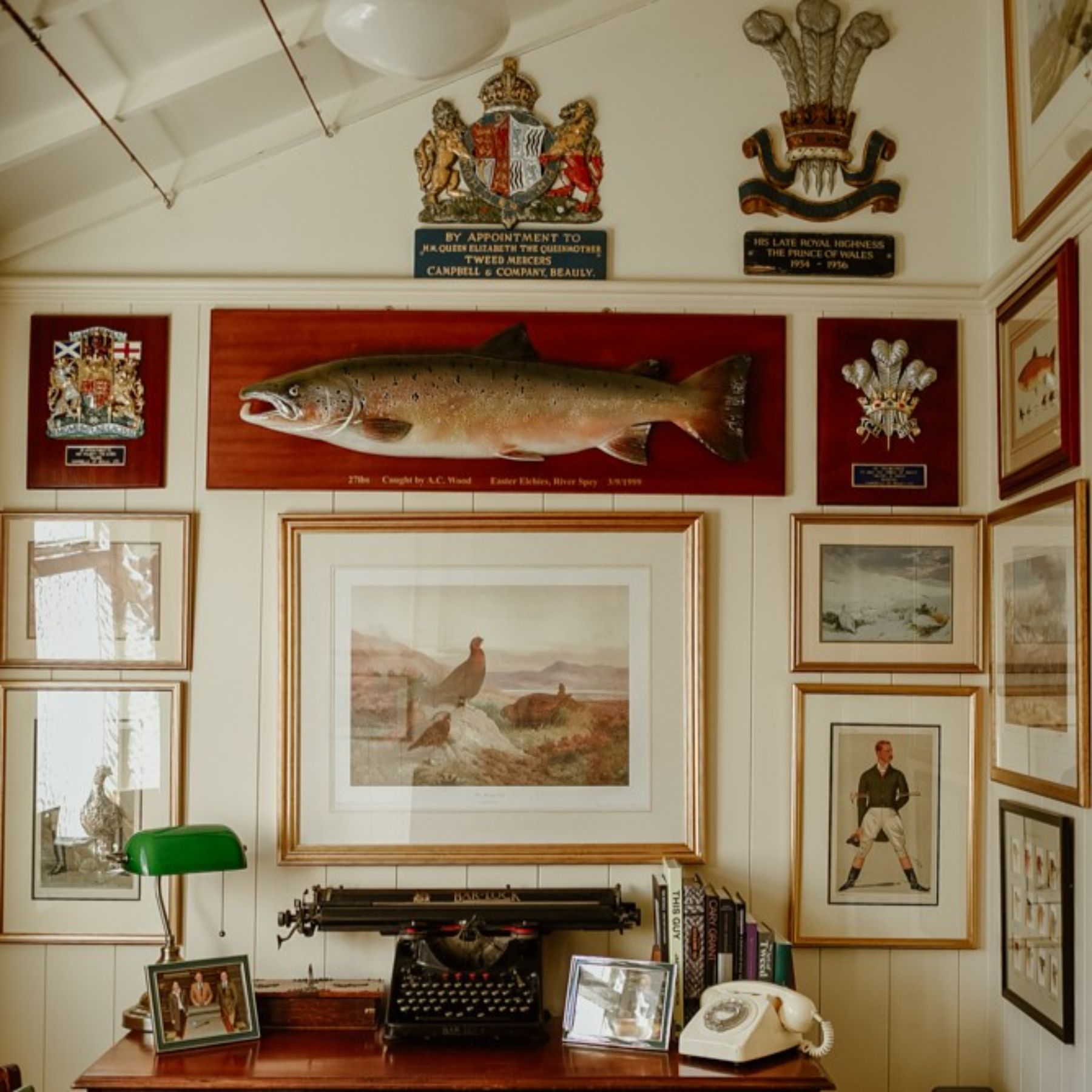 A vintage study with a typewriter, rotary phone, and books on a wooden desk. The wall displays fish, birds, portraits, and royal crests in frames, a mounted fish, and several plaques.