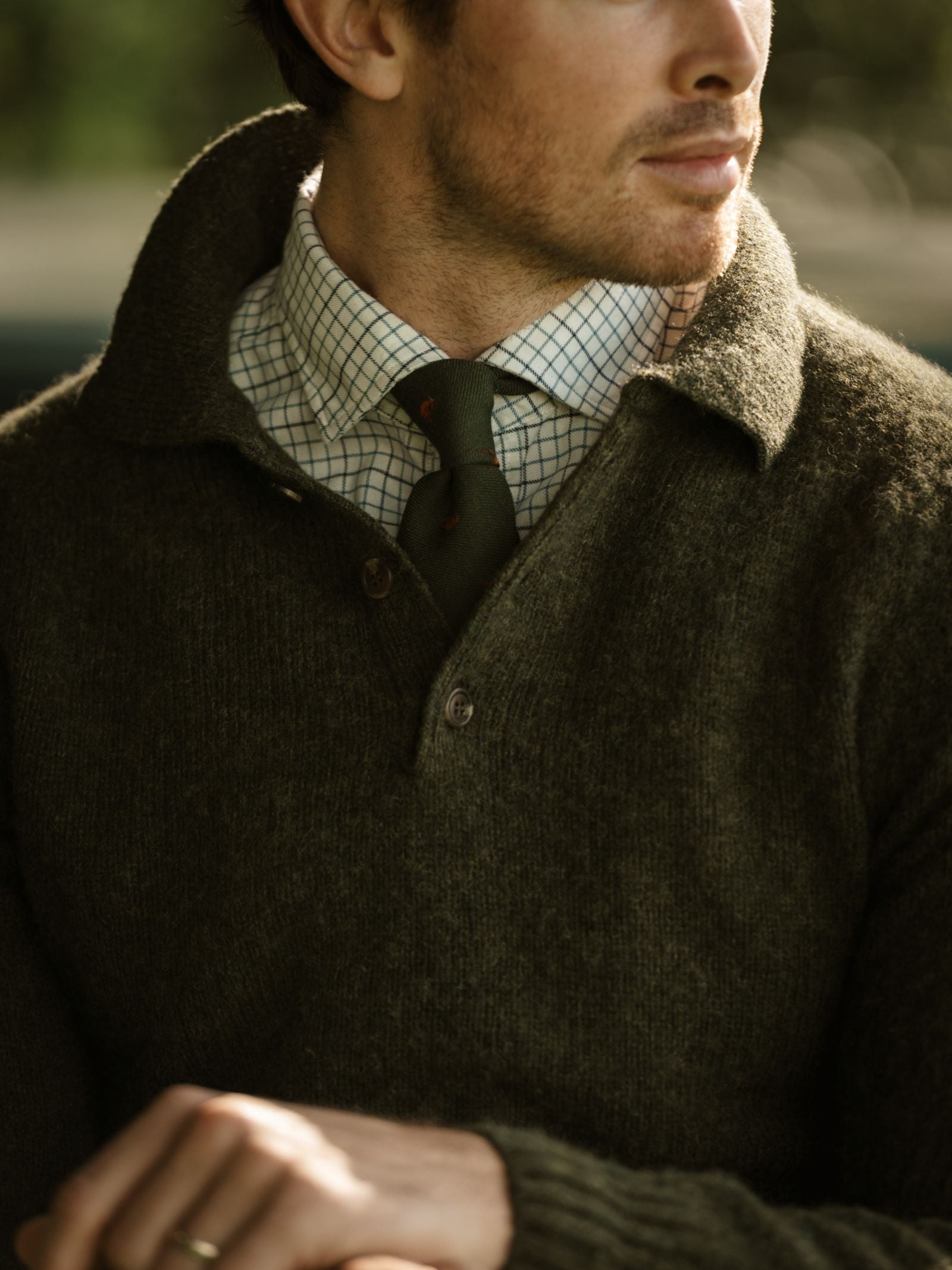 A man wears a Campbell's of Beauly Brushed Shetland Rugby Jumper in olive green over a checkered shirt and dark tie, sitting with his head turned to the side. The image highlights the sweater and upper body details.