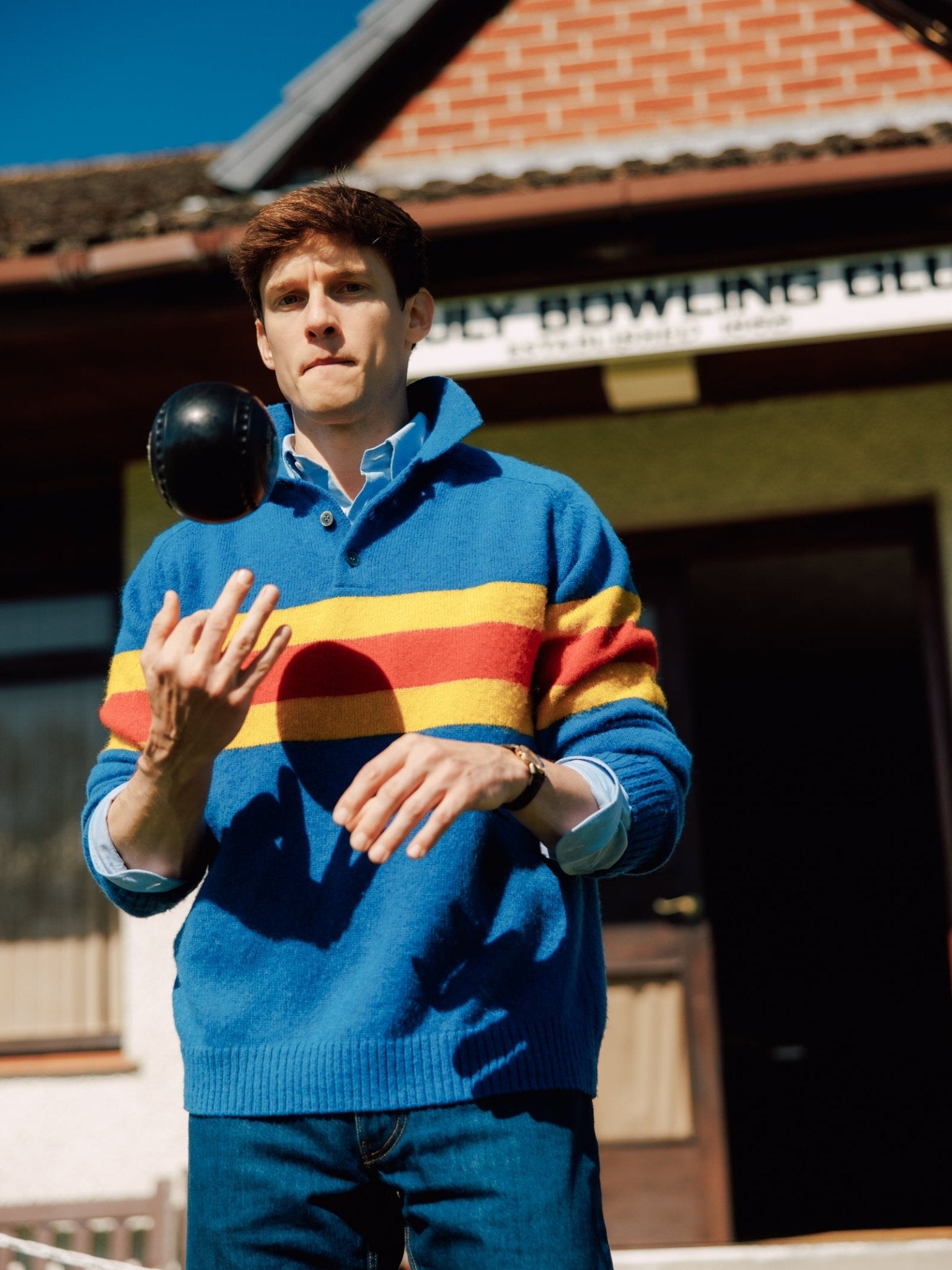 A young man wearing the Campbell's of Beauly Brushed Shetland Stripe Rugby sweater with yellow and red stripes tosses a black ball in the air outside a brick and green building.