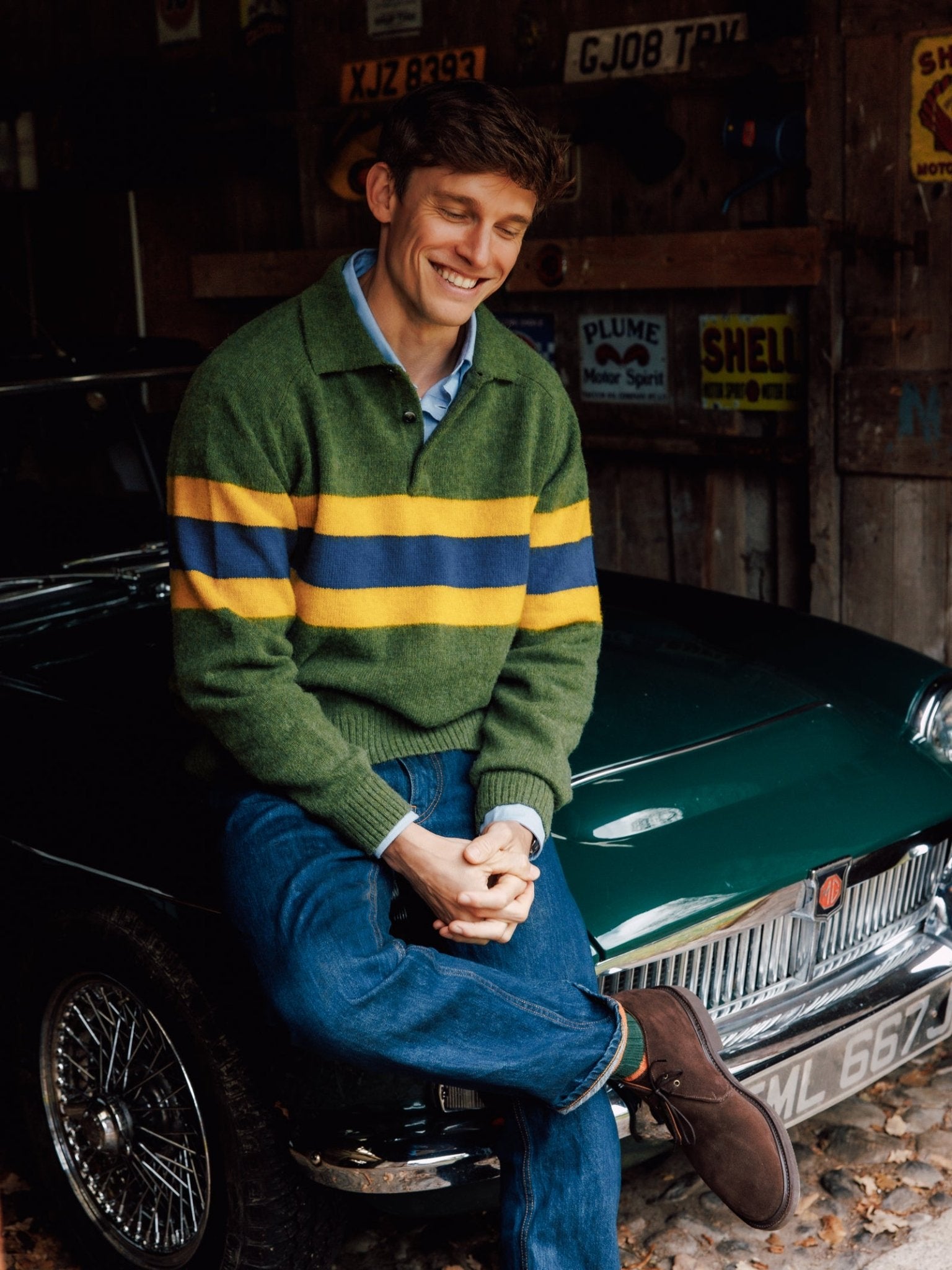 A young man wearing the Campbell's of Beauly Brushed Shetland Stripe Rugby in green smiles while sitting on a classic green car in a rustic garage, vintage signs displayed on the wall behind him.