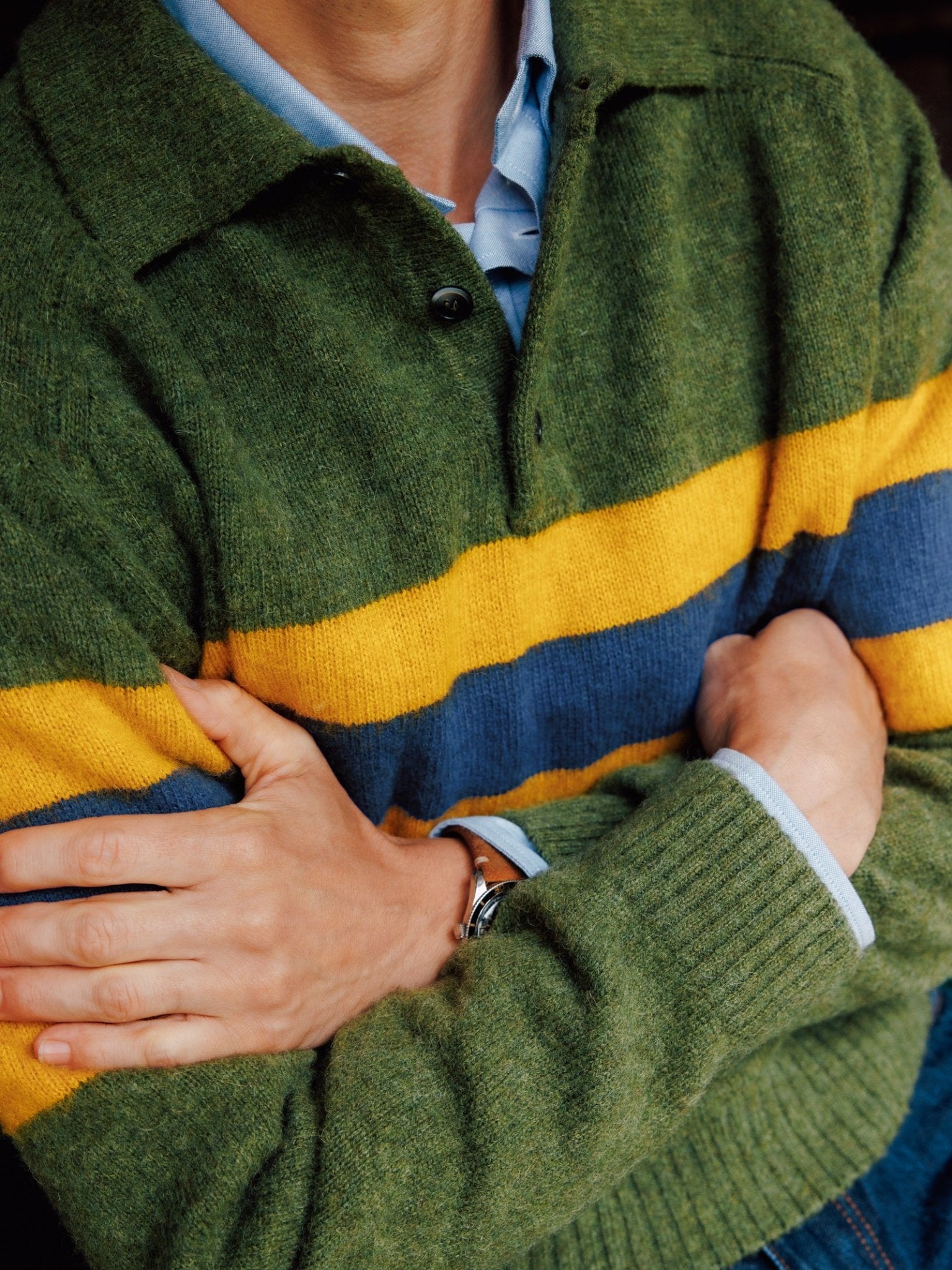 A person folds their arms, wearing the Campbell's of Beauly Brushed Shetland Stripe Rugby sweater. Green with yellow and blue stripes, it’s layered over a light blue shirt. The cuffs show at the wrists, and a watch is worn on the left wrist.