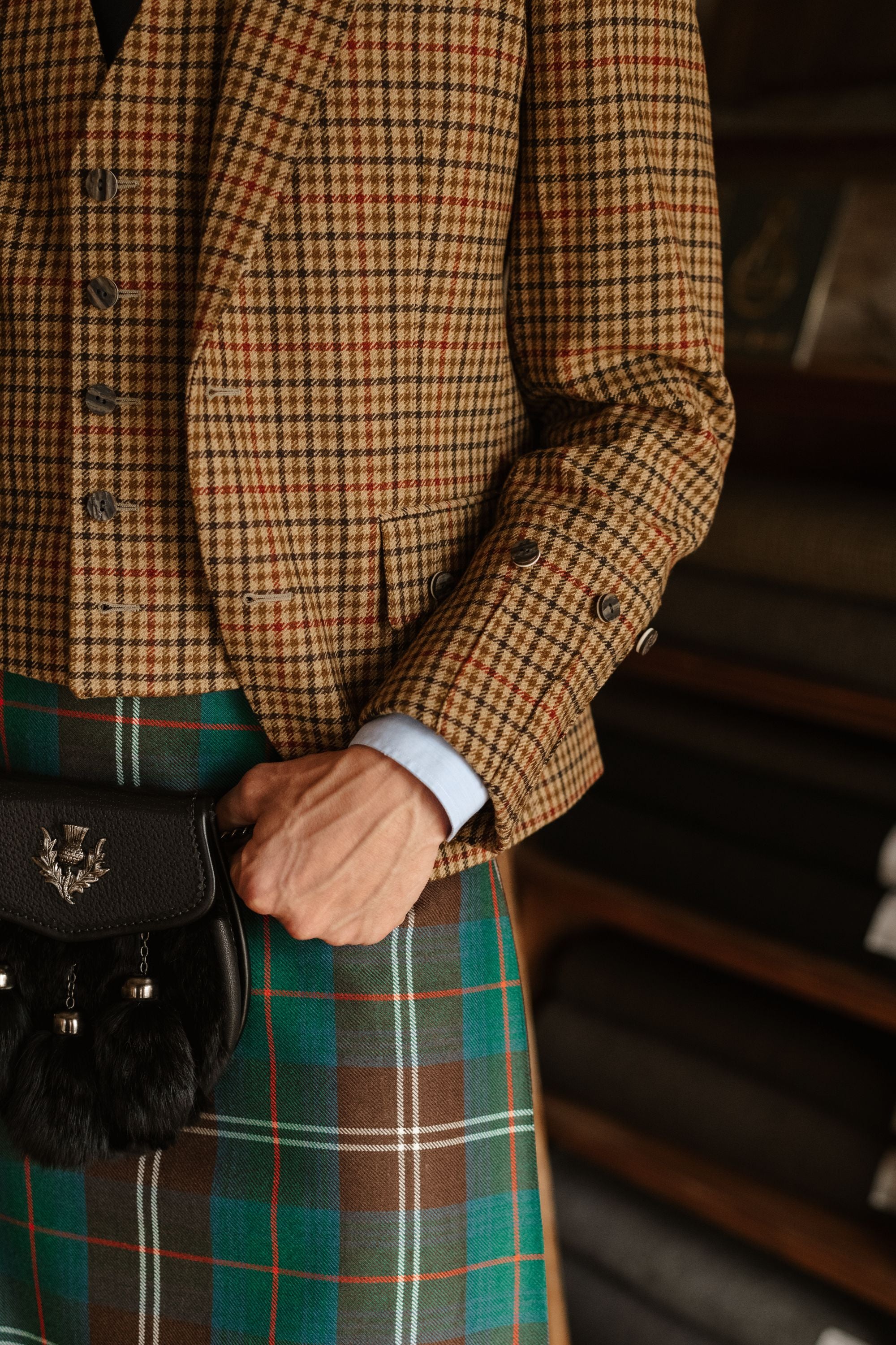 A person wearing a brown plaid jacket and vest, a blue shirt, and a green tartan kilt holds a black sporran with silver detailing. Fabric rolls are visible in the background.