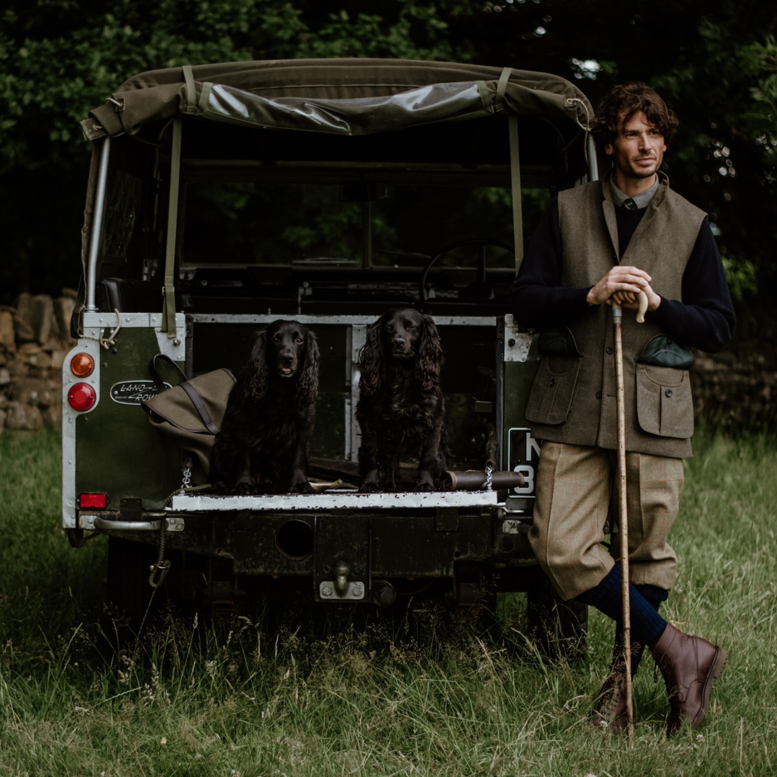 A man in country attire leans on a walking stick beside an open Land Rover, with two black spaniel dogs sitting on the tailgate in a grassy, rural setting.