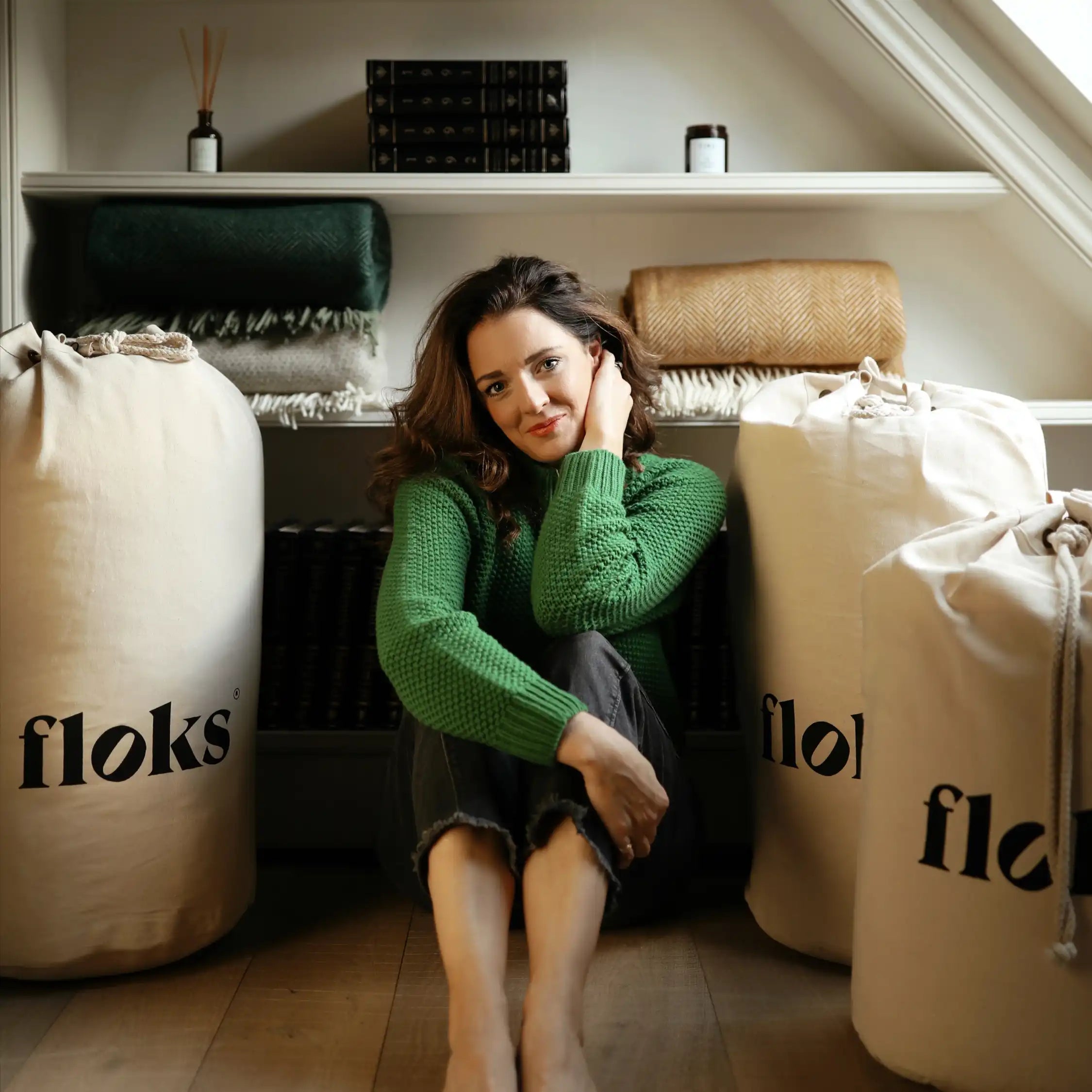 A woman in a green sweater sits barefoot on the floor between large canvas bags labeled floks, with folded blankets and books on shelves behind her in a cozy, attic-like room.