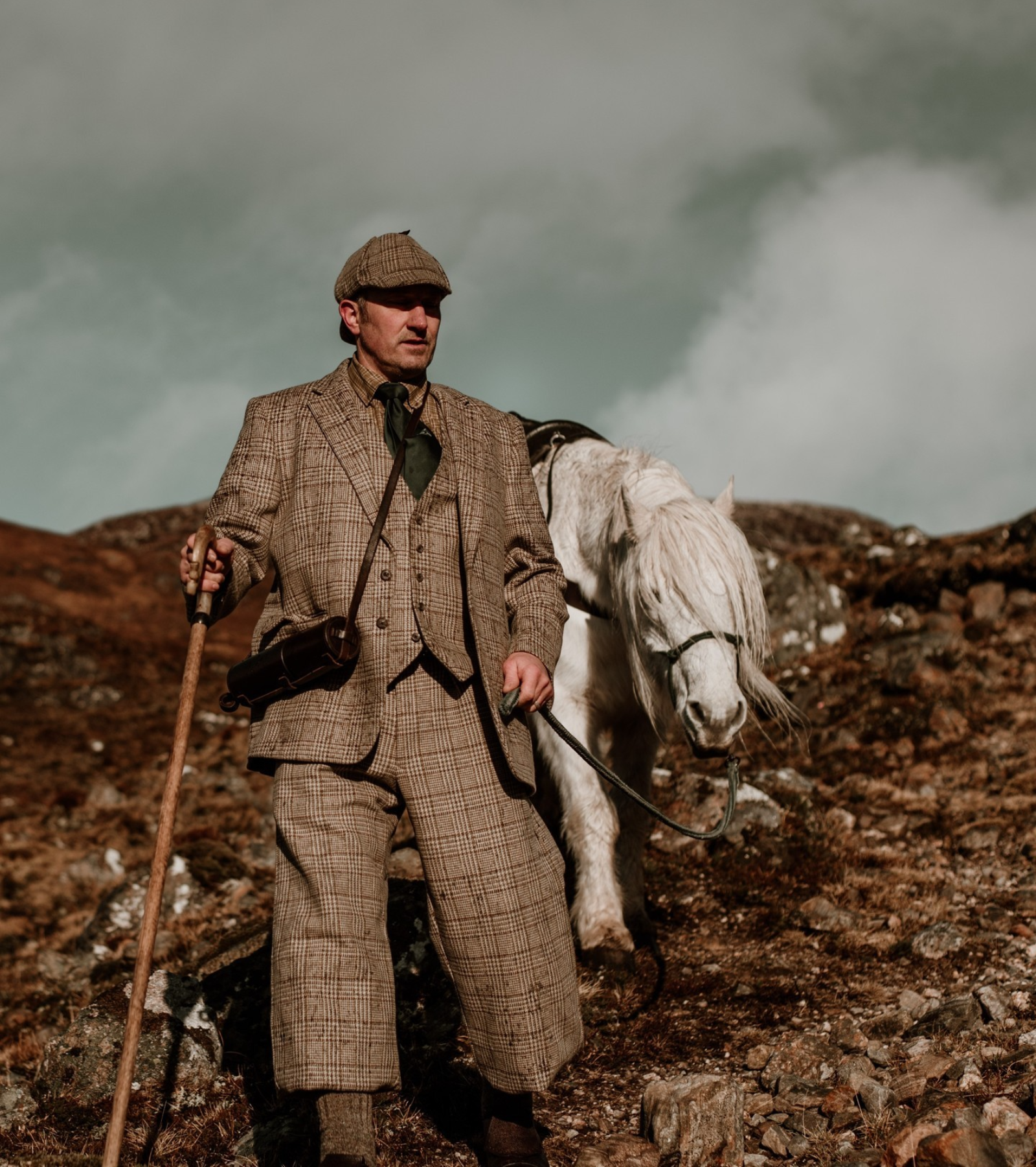 A man in vintage tweed hunting attire walks on rocky terrain with a wooden walking stick, leading a white horse behind him. The sky is cloudy and the landscape is rugged and natural.