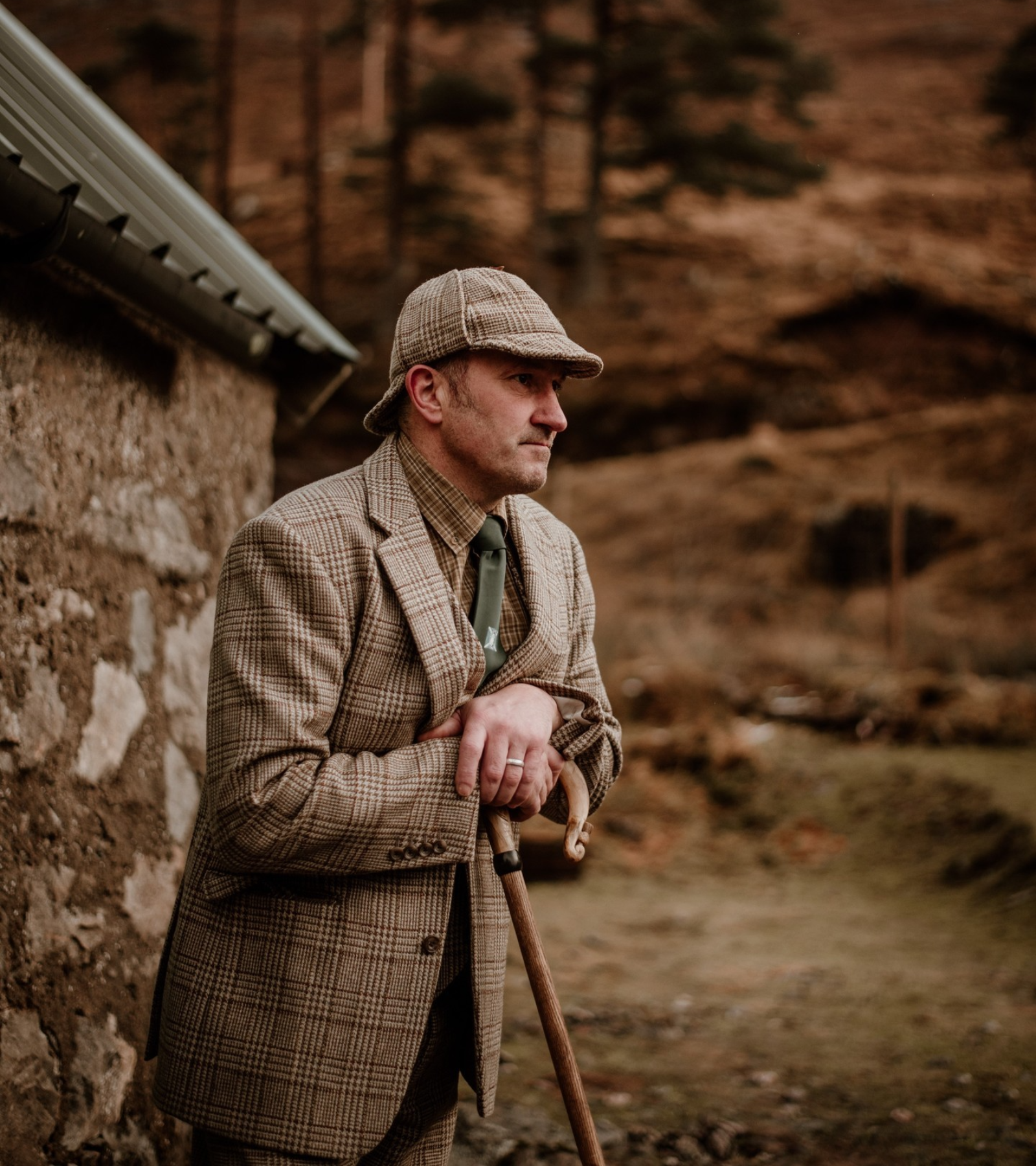 A man in a plaid suit, tie, and matching flat cap stands outdoors beside a stone building, leaning on a wooden cane, with a background of trees and brown, hilly terrain.