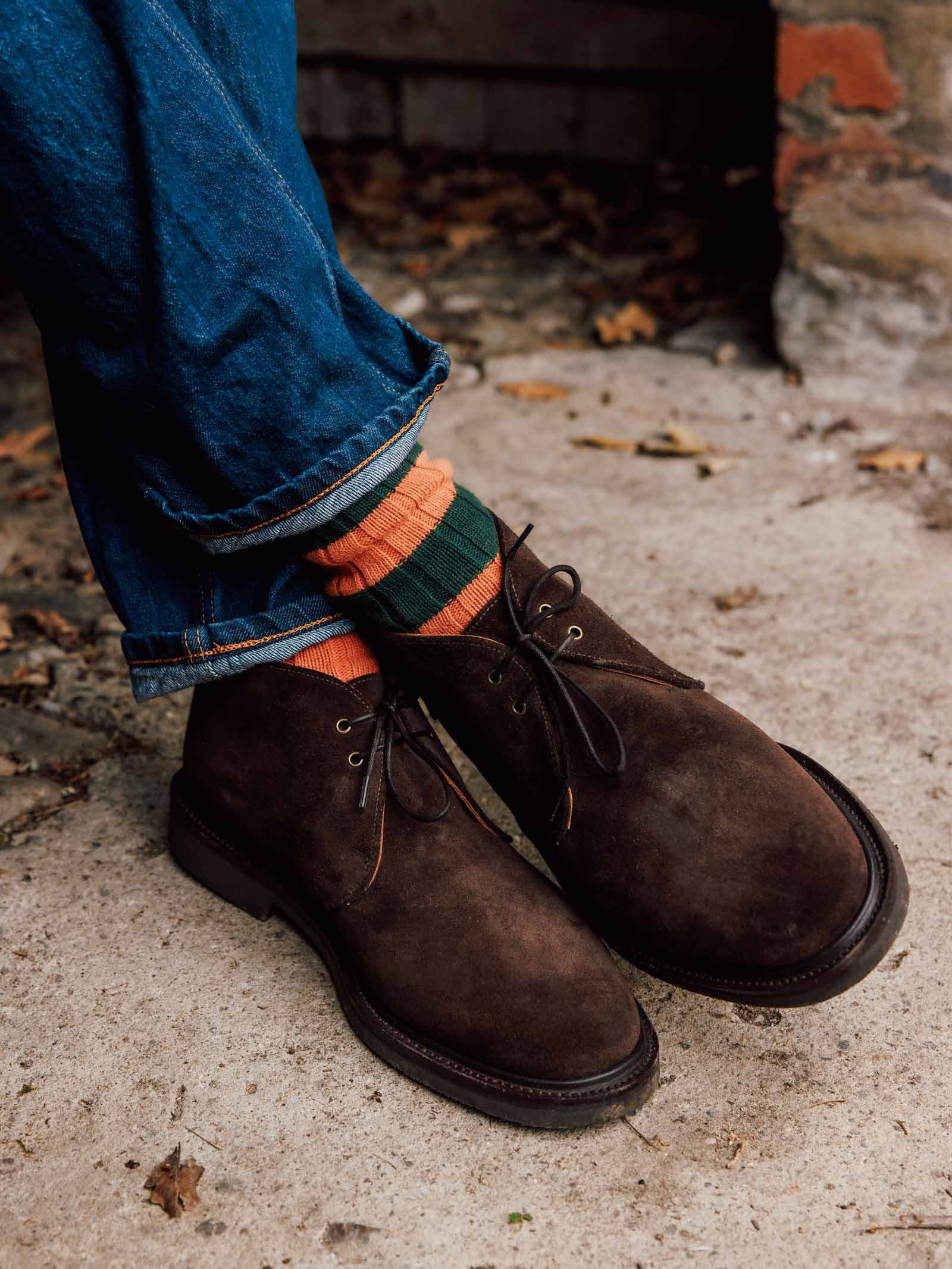 A person is wearing Lomond Boots by Campbell’s of Beauly in chocolate brown suede, paired with blue jeans and orange-and-green striped socks, standing on a concrete surface scattered with leaves and a partially visible brick wall in the background. The Goodyear welted construction of the Lomond Boots adds both durability and classic style to the ensemble.