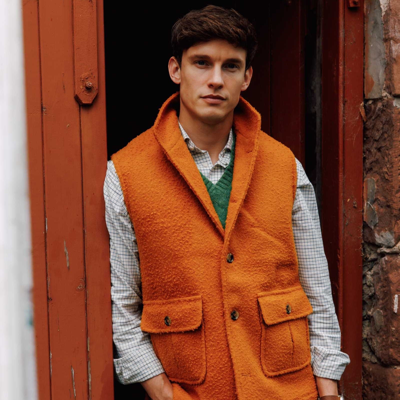 A young man with short brown hair stands in a doorway, wearing a checked shirt, a green sweater vest, and an orange wool vest with large pockets. He looks calmly at the camera. The doorway and wall are reddish-brown.