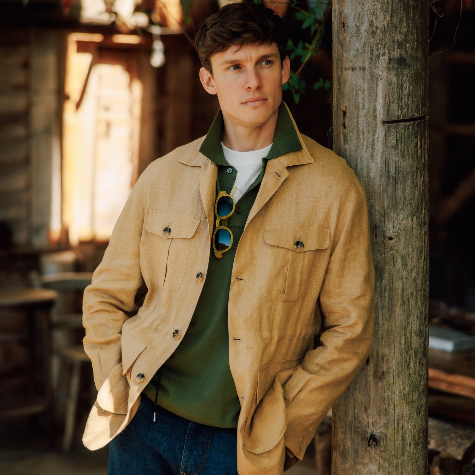 A young man with short brown hair leans against a wooden post, wearing a tan jacket over a green collared shirt with sunglasses hanging from the collar, in a rustic, sunlit setting.