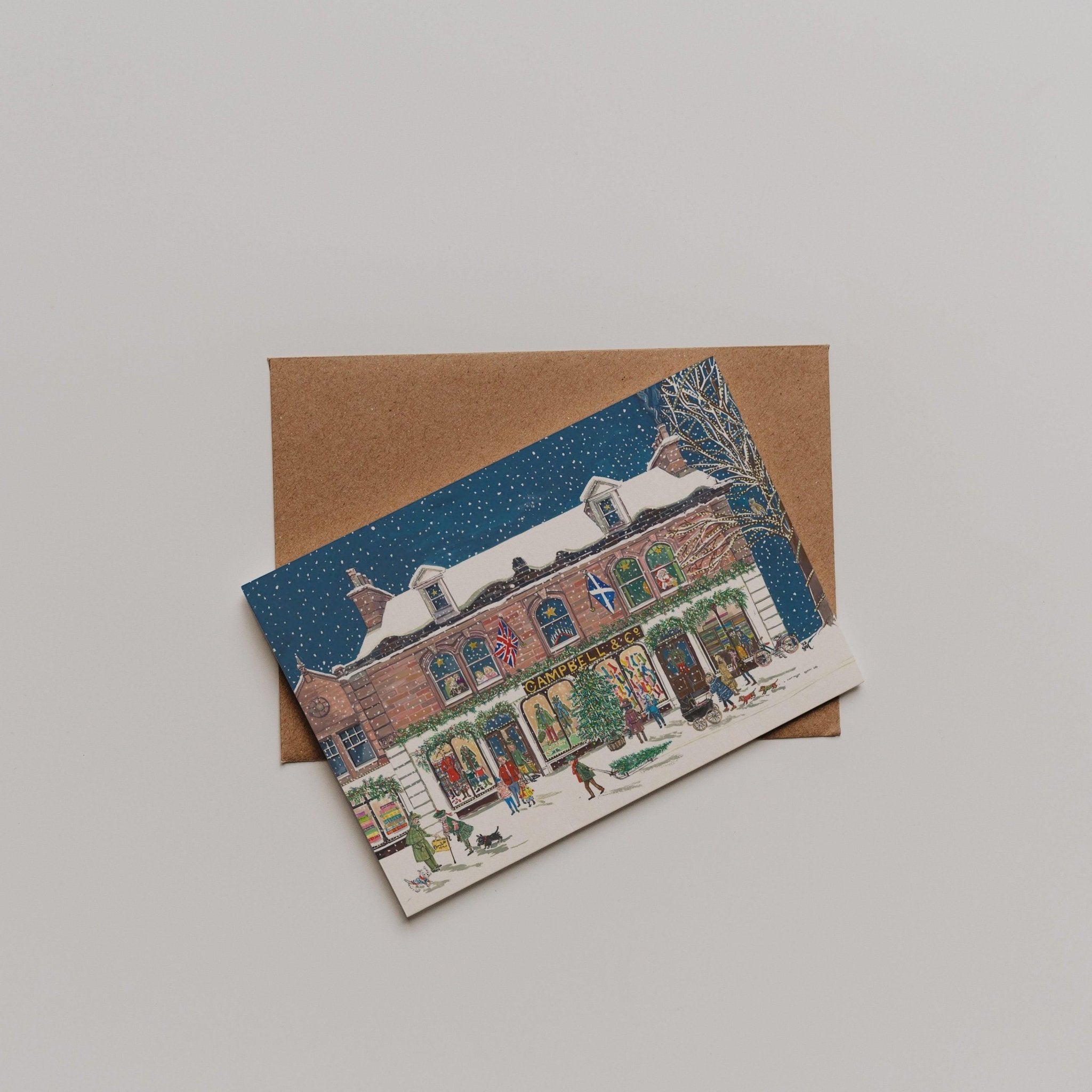 The Campbells at Christmas Greeting Card by Campbells of Beauly displays a snowy winter scene with festive buildings and figures, set above a brown envelope—an ideal thank you card for any occasion.