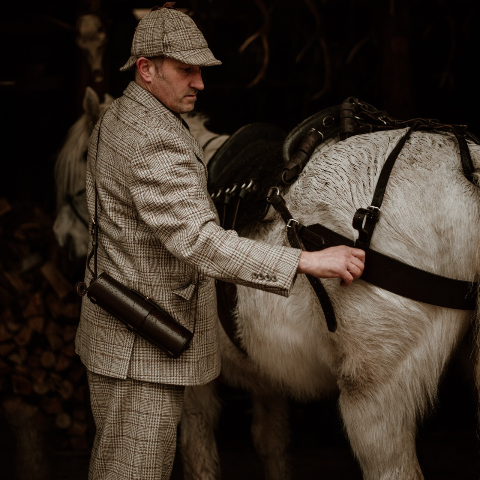 A man in a plaid suit and hat adjusts the harness on a white horse. He carries a cylindrical case over his shoulder. The background is dimly lit with stacked firewood visible.