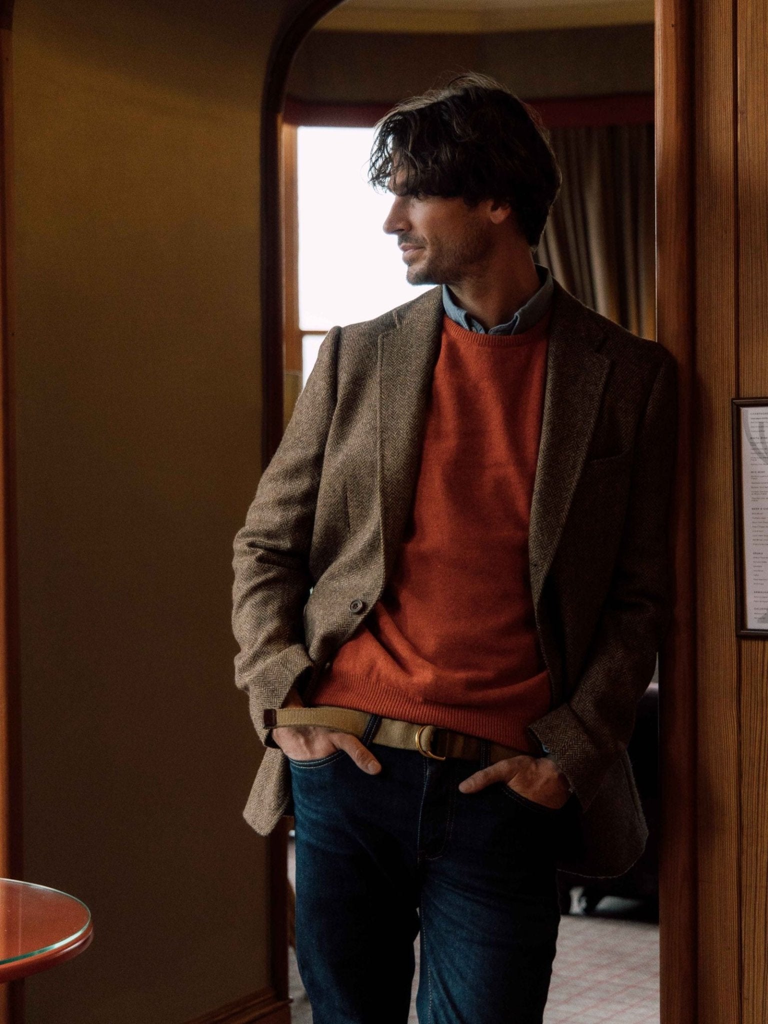 A man with medium-length hair stands indoors by a wooden door, wearing a brown blazer over the Campbell's of Beauly Lambswool Crew Neck Jumper and blue jeans, hands in his pockets, looking to the side.