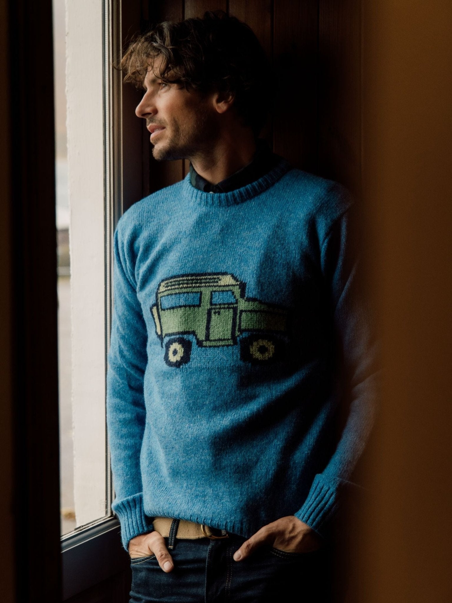 A man with wavy brown hair stands by a window, wearing the Campbell's of Beauly Unisex Land Rover Defender Jumper in Blue Lovat. Featuring a green car motif, the classic knitwear is softly illuminated by natural light as he gazes outside.