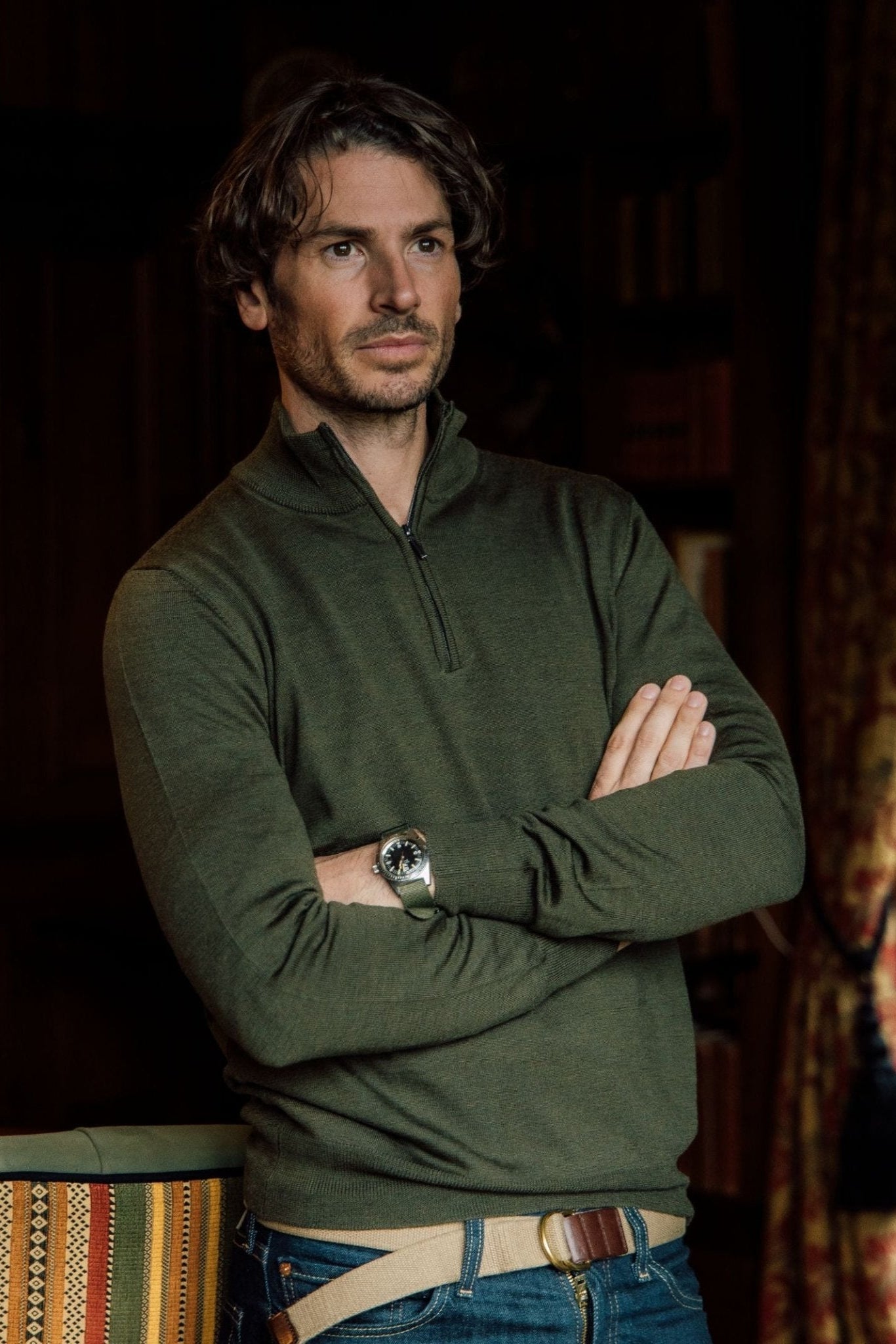 A man with wavy brown hair, wearing blue jeans and a green Campbell's of Beauly Auld Stock Merino Quarter Zip Jumper, stands with arms crossed and a wristwatch. He looks thoughtful in a softly lit room with shelves and a patterned curtain behind him.