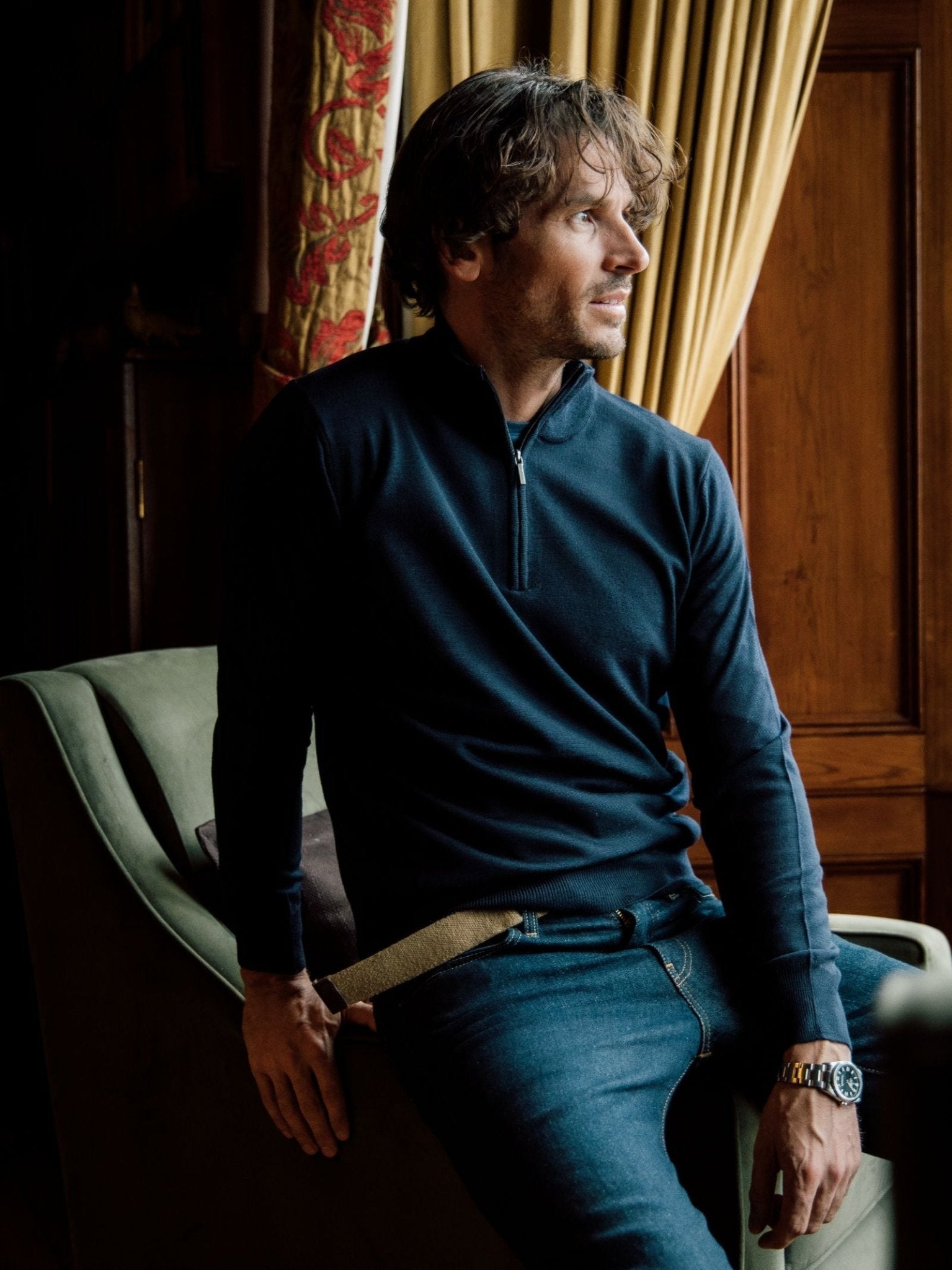 A man with wavy hair, wearing the Campbell's of Beauly Auld Stock Merino Quarter Zip Jumper and jeans, sits on a chair arm in a wood-paneled room with gold curtains, gazing thoughtfully to the side.