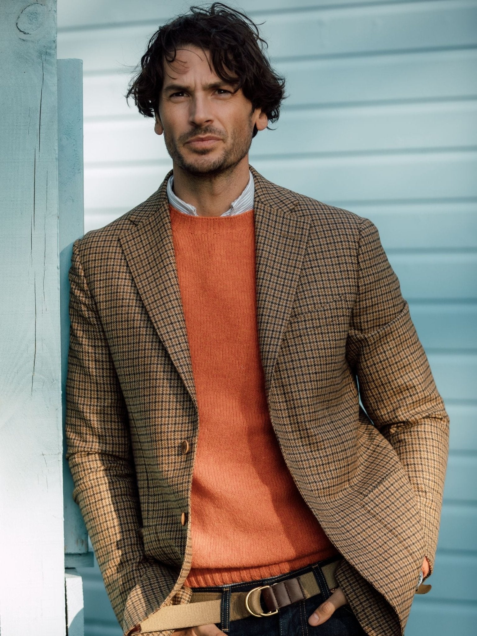 A man with wavy brown hair wears the Campbell's of Beauly Auld Stock Gun Club Jacket over an orange sweater, a light blue shirt, and jeans. He stands against a light blue wooden wall, one hand in his pocket, looking confidently at the camera.