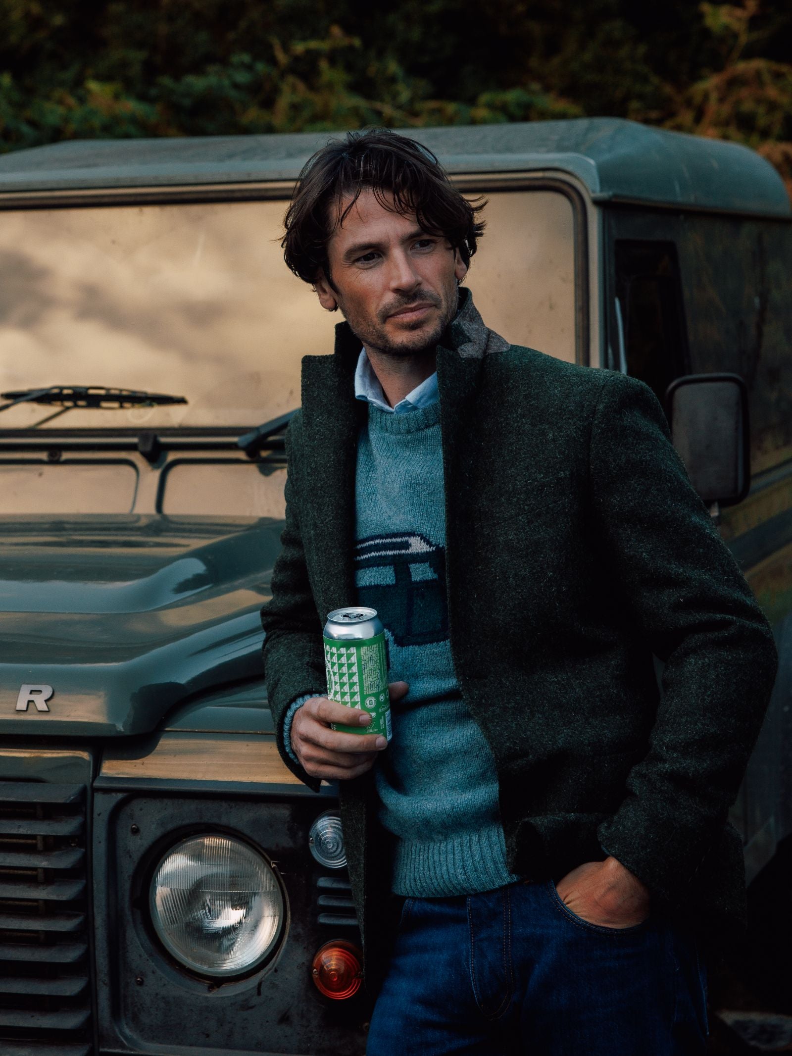 A man with wavy brown hair wears the Campbell’s of Beauly Unisex Land Rover Defender Jumper and a green jacket, standing by a green Land Rover with a can in hand, evoking timeless British motoring style.