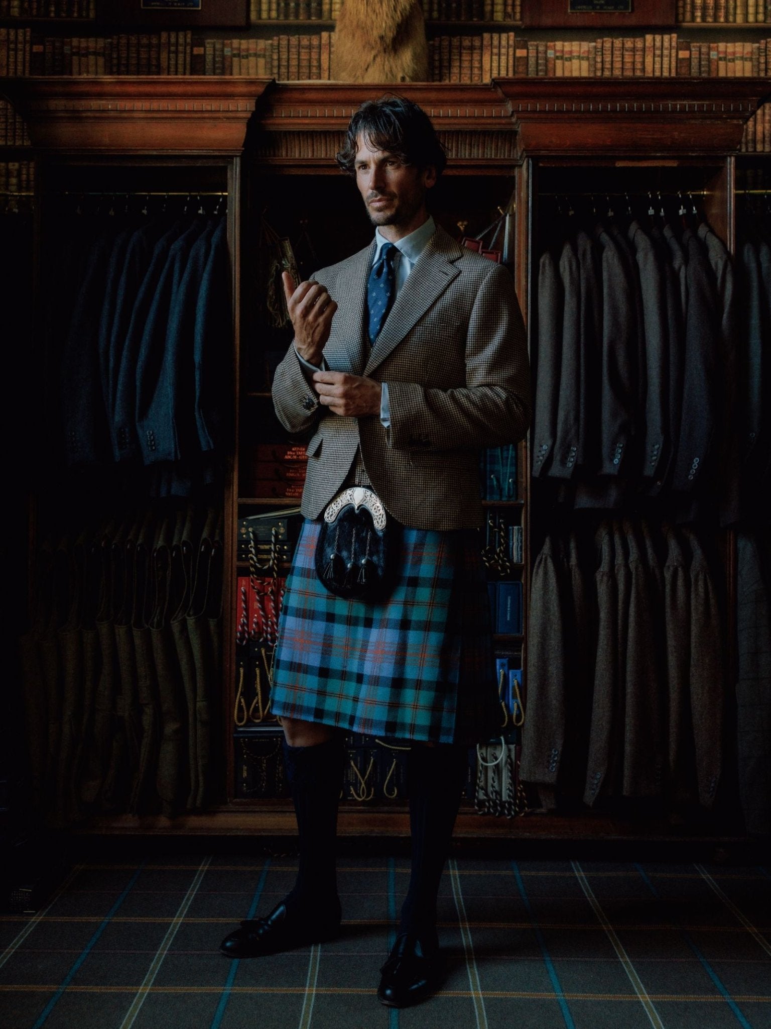 A man in Highland dress stands before shelves of suits, adjusting his cuff in an elegant room. He wears a brown jacket, blue tartan kilt, dress shoes, a sporran, and Campbell’s of Beauly Kilt Socks.