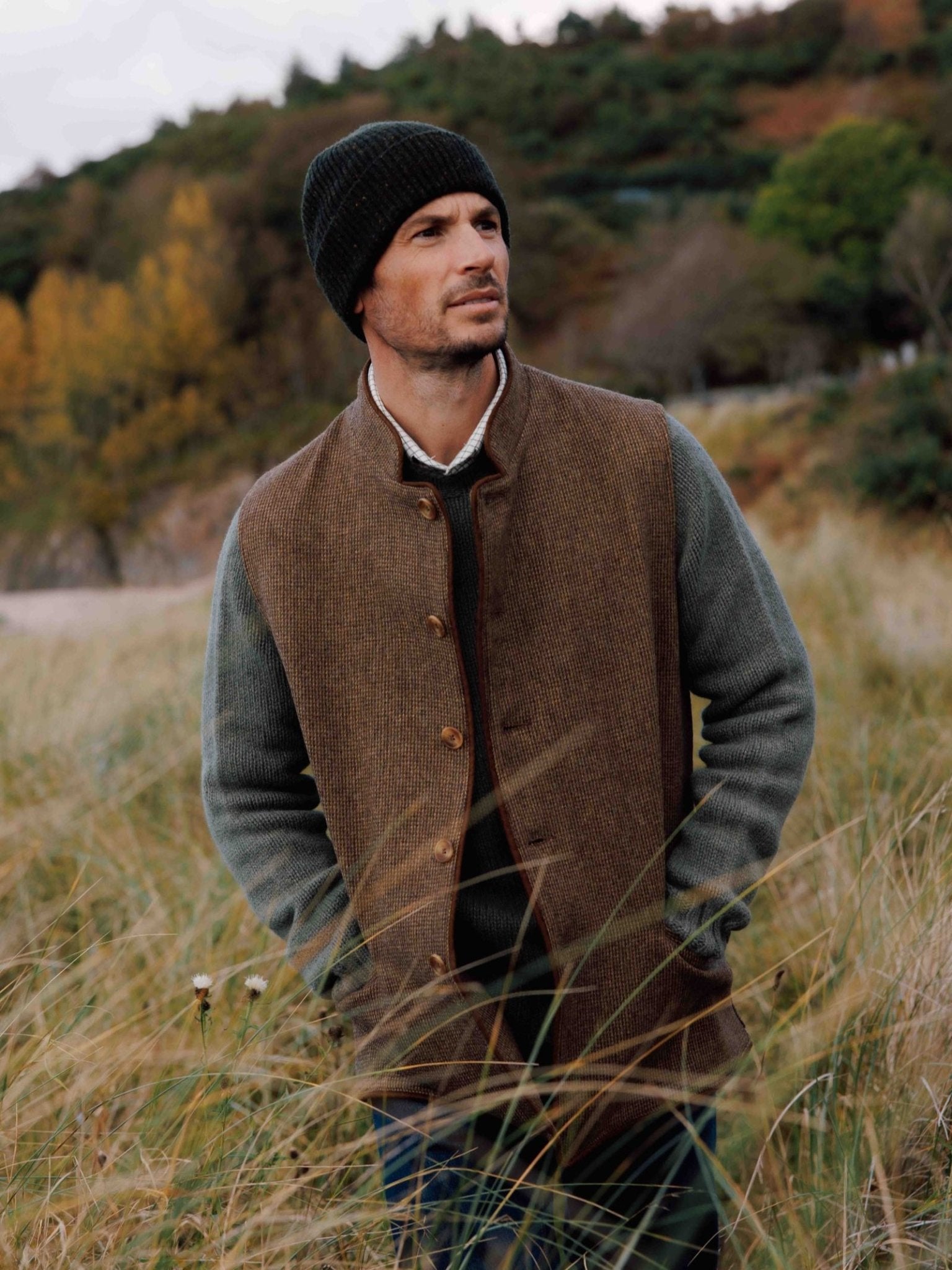 A man wearing the Campbell's of Beauly Tweed Nehru Gilet, a green sweater, and a black knit beanie stands in tall grass, gazing into the distance with autumn trees and a cloudy sky behind him.