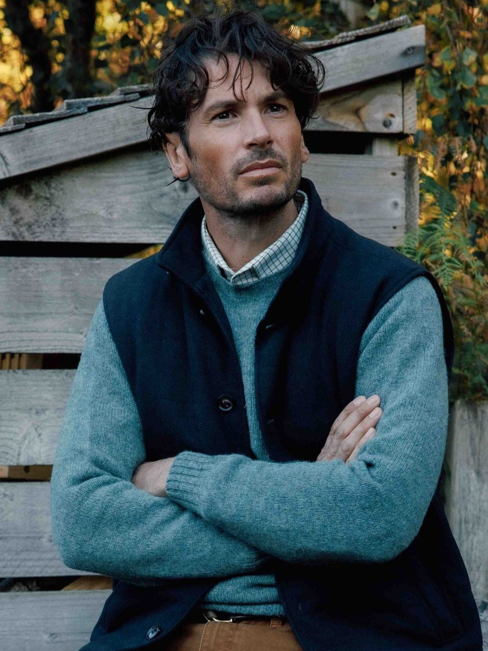 A man with curly dark hair, dressed in a blue sweater over a checkered shirt and layered with the Campbell’s of Beauly Quilted Buttoned Gilet, sits outdoors with his arms crossed in front of a rustic wooden structure.