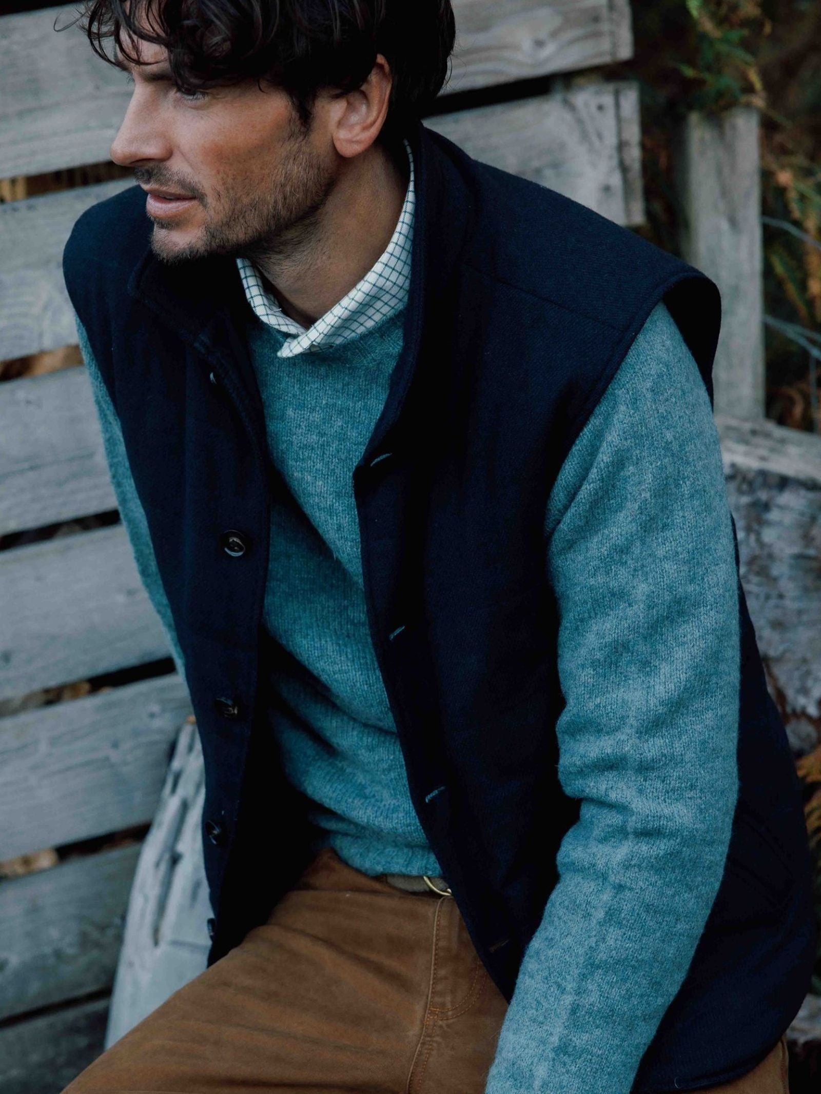 A man with dark hair sits on a wooden bench, wearing a blue sweater layered over a collared shirt, paired with the Campbell’s of Beauly Quilted Buttoned Gilet in navy tartan and brown pants. He gazes thoughtfully to the side.