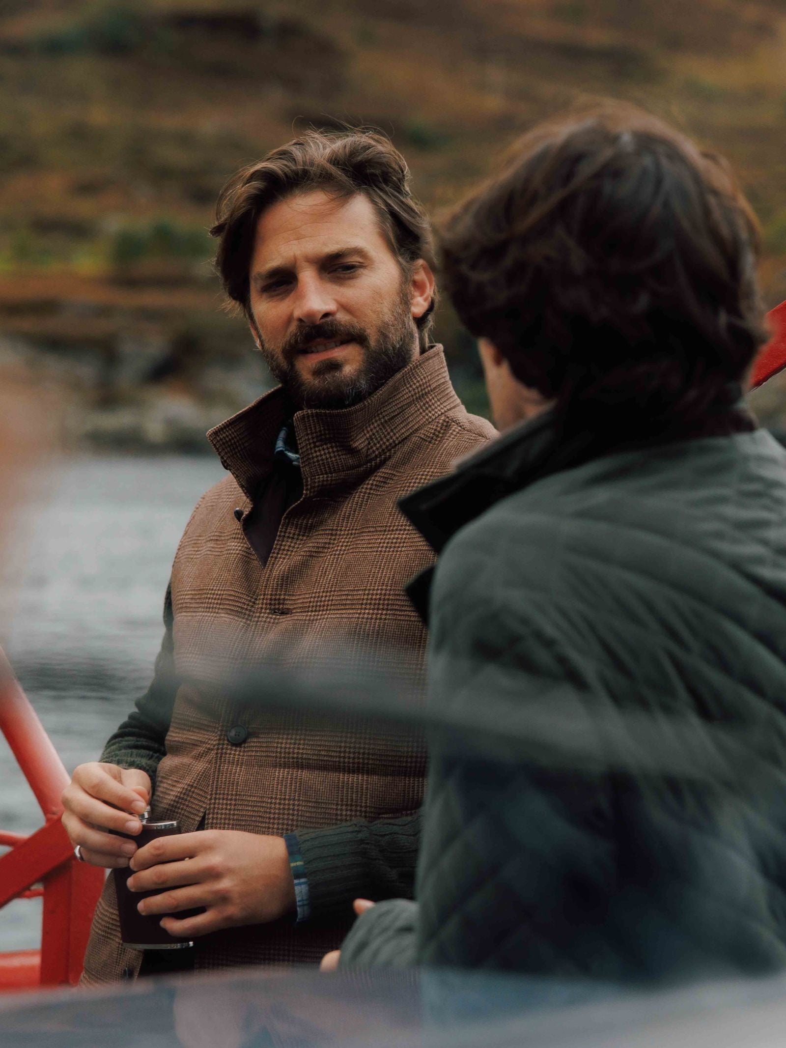 Two men chat by the water. One, drink in hand, wears a British tweed jacket; the other, seen from behind, dons a Campbell's of Beauly Cashmere Gun Club Quilted Gilet—timeless additions that elevate their country style against a relaxed backdrop.