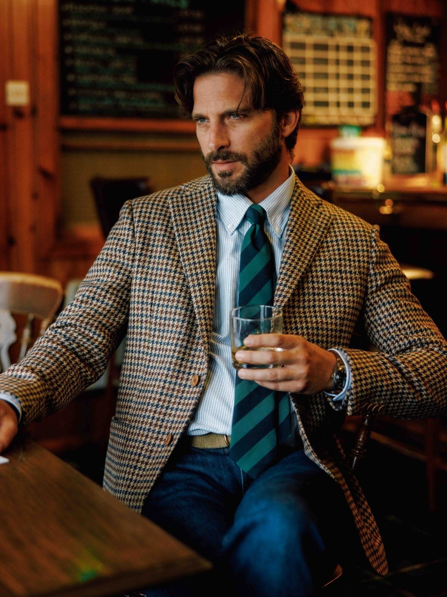 A man with wavy brown hair and a beard sits at a wooden bar table, wearing a checked blazer and the Green & Navy Stripe Tie by Campbell's of Beauly, holding a whiskey glass with chalkboard menus in the background.