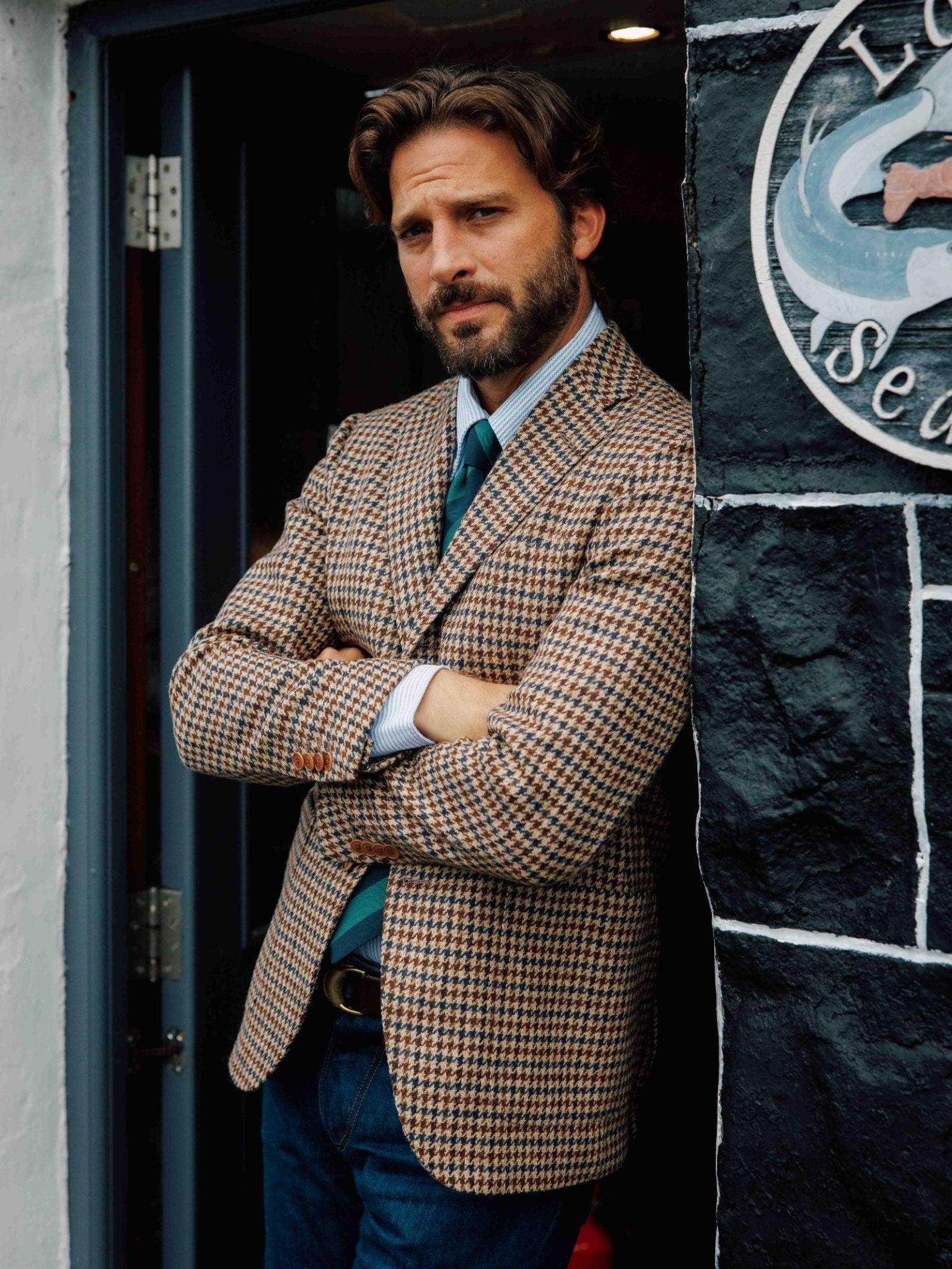 A bearded man with wavy brown hair stands in a doorway, arms crossed, wearing a checked blazer, striped shirt, Campbell's of Beauly Green & Navy Stripe Tie, and jeans. A circular fish sign is partially visible on the wall beside him.