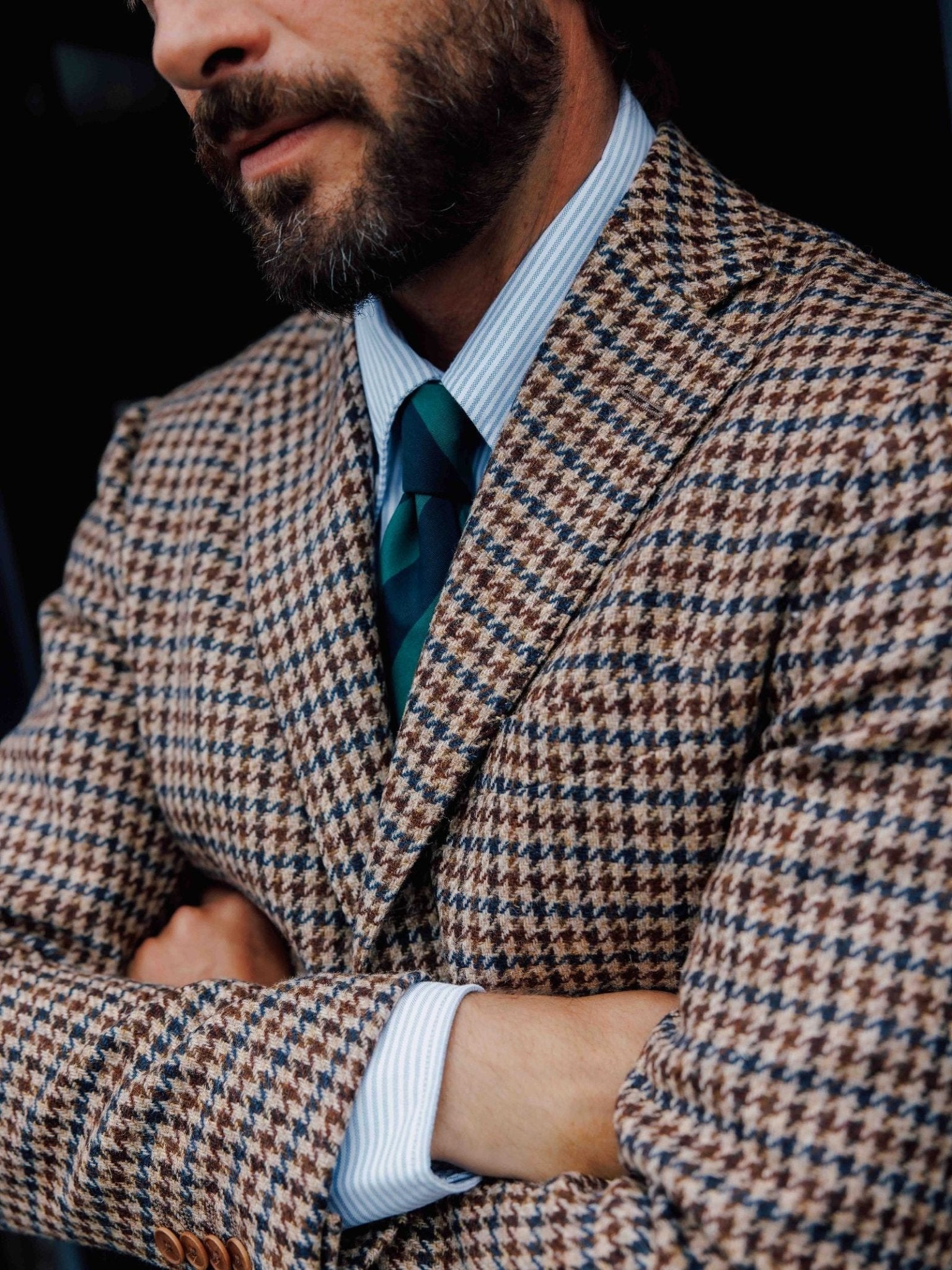 A man with a beard crosses his arms while wearing a brown and blue houndstooth suit, a light blue striped shirt, and the Green & Navy Stripe Tie by Campbell's of Beauly for timeless style.