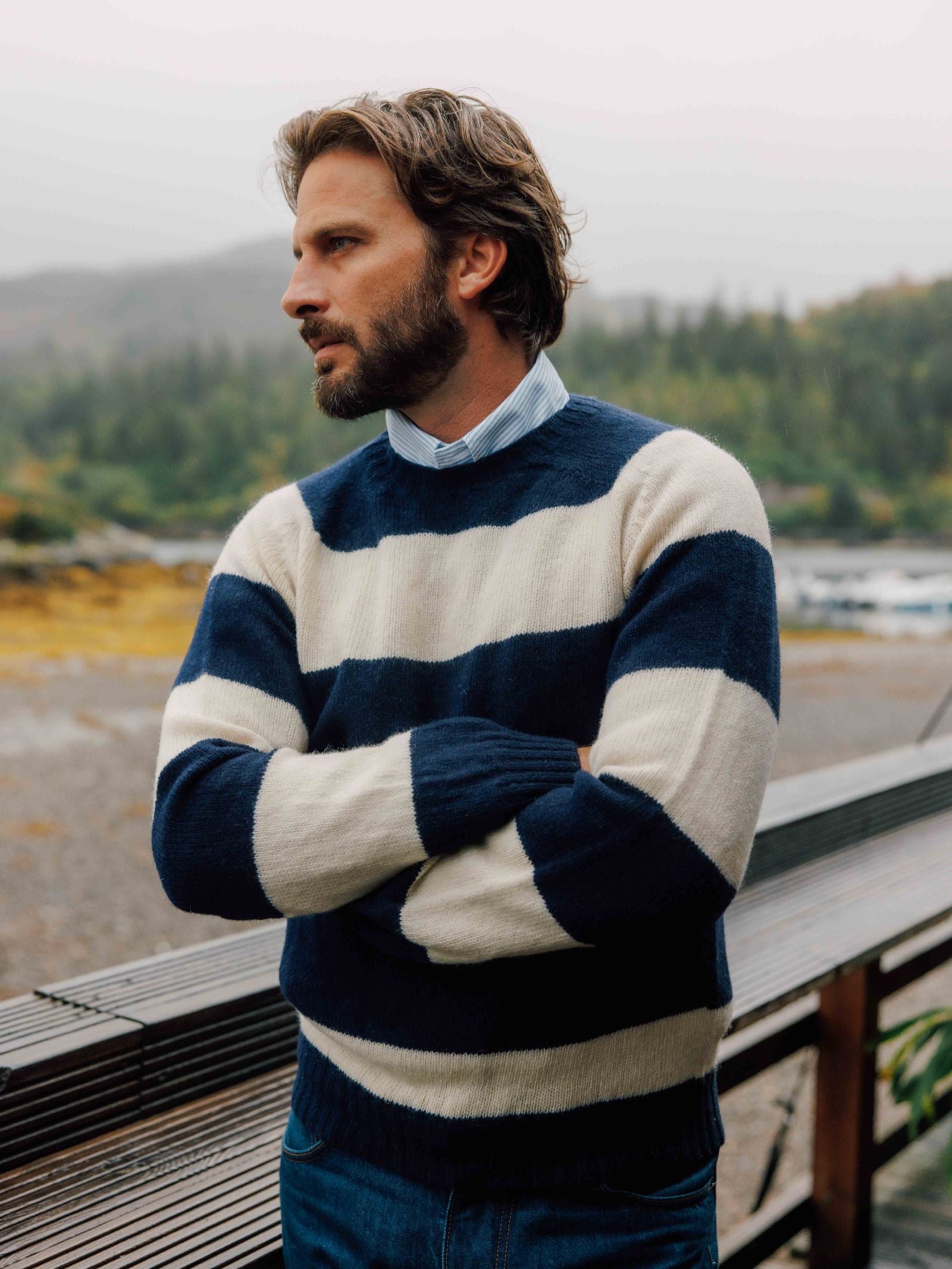 A man with medium-length brown hair and a beard stands by a wooden railing outdoors, wearing Campbell's of Beauly Auld Stock Shetland Stripe Crew Jumper in blue-and-white over a collared shirt, with a blurred forest and water behind him.