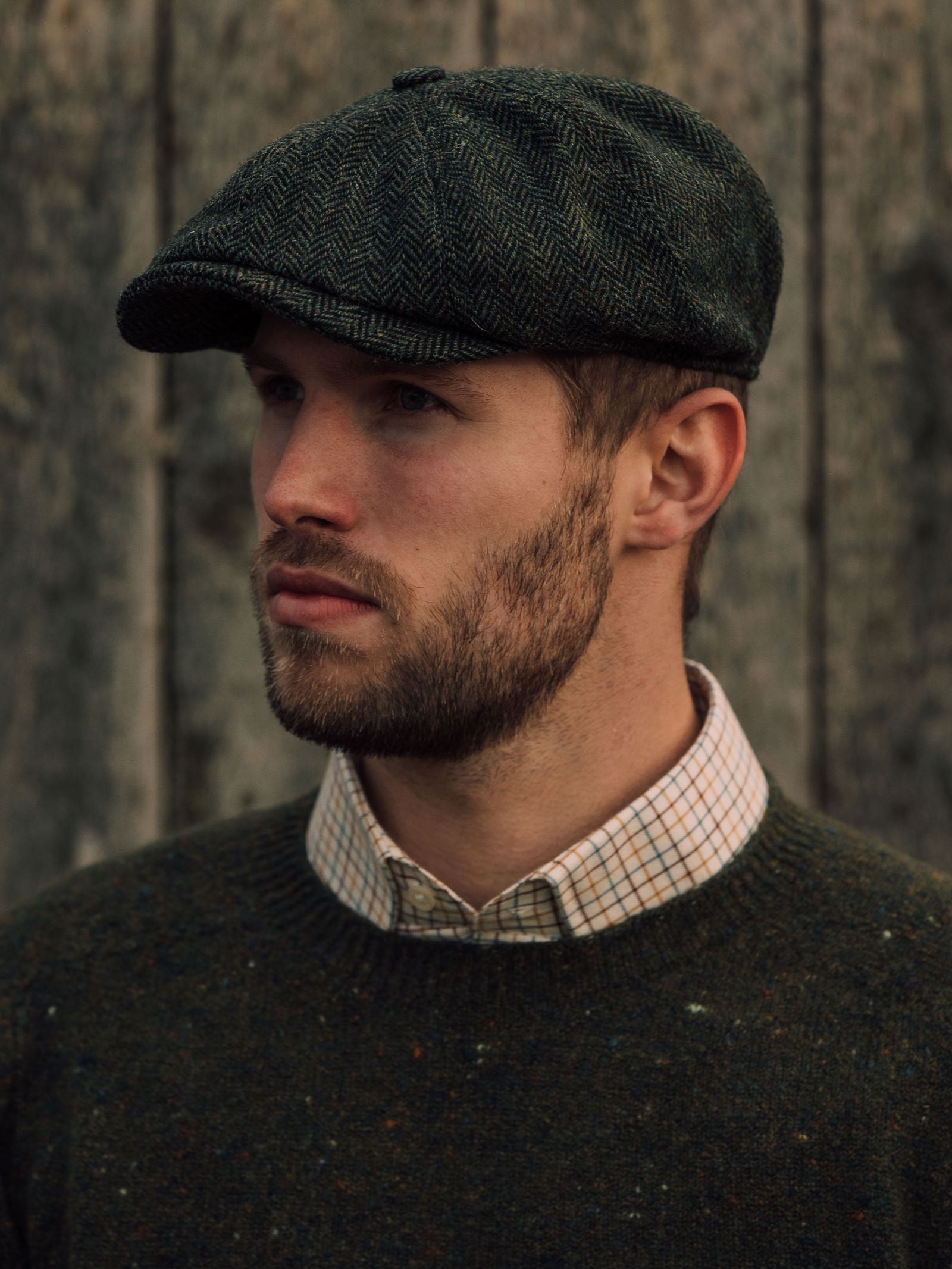 A man with a short beard wears the Campbells of Beauly Farrar Cap, paired with a green sweater and checked shirt, standing by a weathered wooden wall and reflecting classic British country heritage.