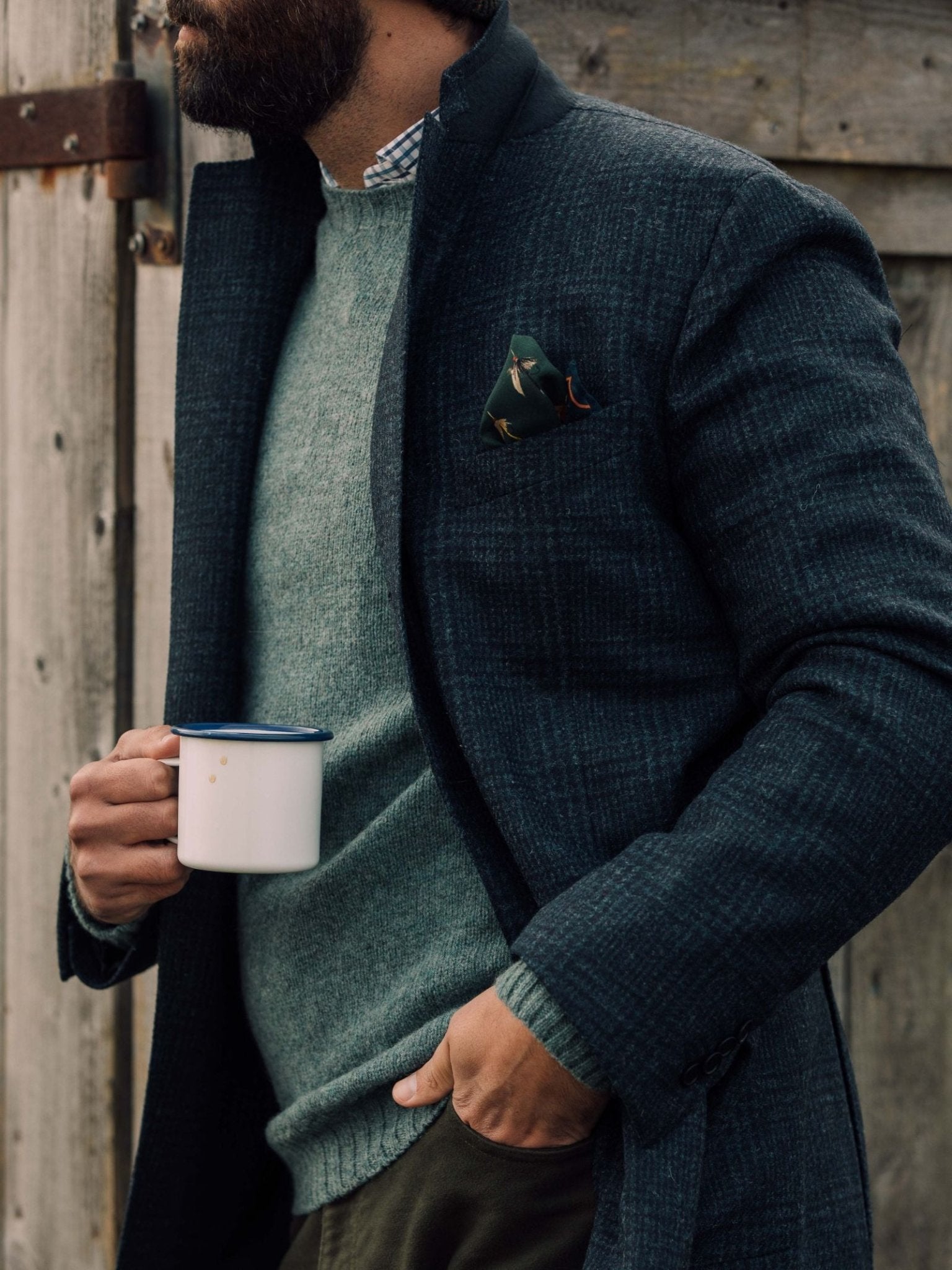 A bearded man holds a white mug with a blue rim. He wears the Auld Stock Glen Check Jacket by Campbell's of Beauly over a green sweater and collared shirt, accented with a patterned pocket square. A rustic wooden door is behind him.