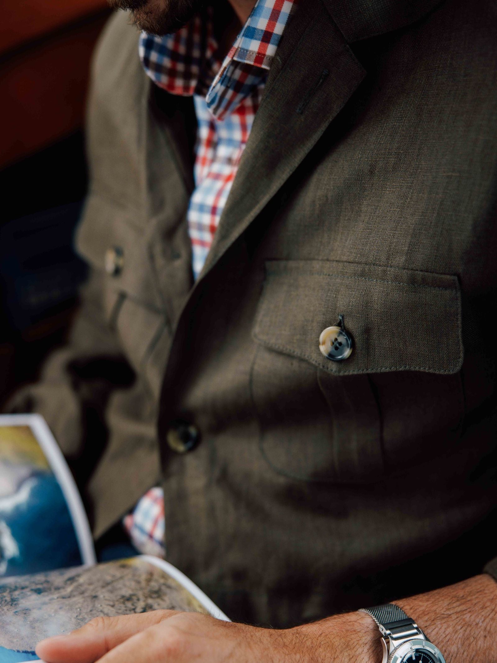 A man in a Campbell's of Beauly Linen Safari Jacket layered over a red, blue, and white plaid shirt holds an open book or magazine. His watch and trimmed beard are partially visible.