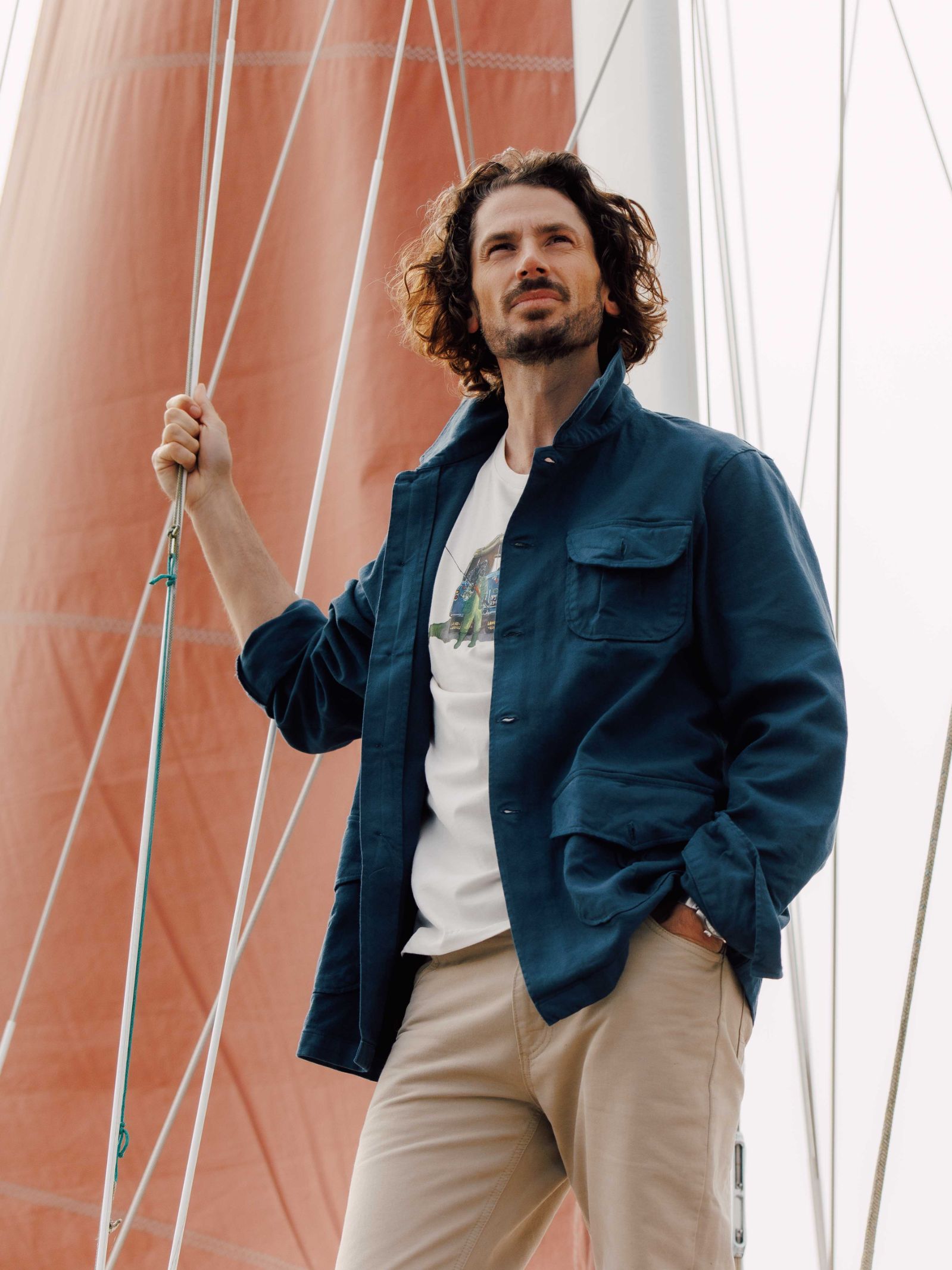 A man with wavy hair wears the blue Auld Stock Field Chore Jacket by Campbell's of Beauly, a white t-shirt, and beige pants as he stands on a sailboat holding a rope, gazing into the distance with a sail behind him.