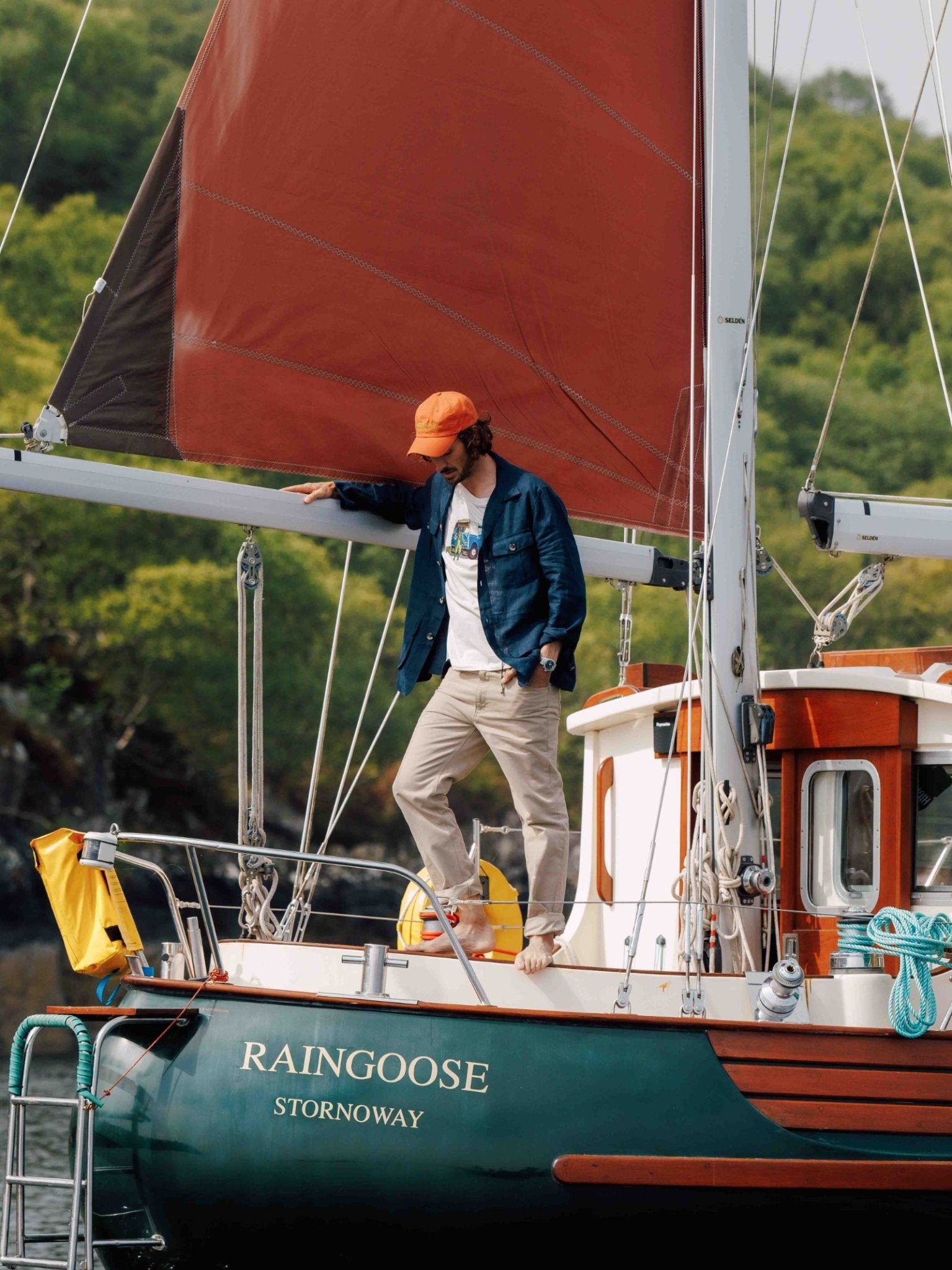A man in an orange cap and a Campbell's of Beauly Auld Stock Field Chore Jacket stands barefoot on the deck of the green and white sailboat “RAINGOOSE STORNOWAY,” with red sails and lush green shoreline behind him.