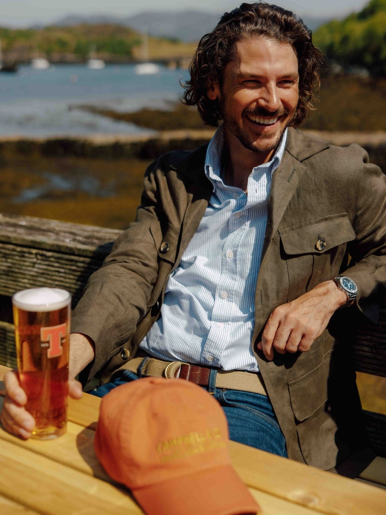 A smiling man with curly hair sits outdoors, holding a pint of beer. He wears the Campbell's of Beauly Linen Safari Jacket over a blue shirt, a watch on his left wrist, and an orange cap rests on the wooden table.